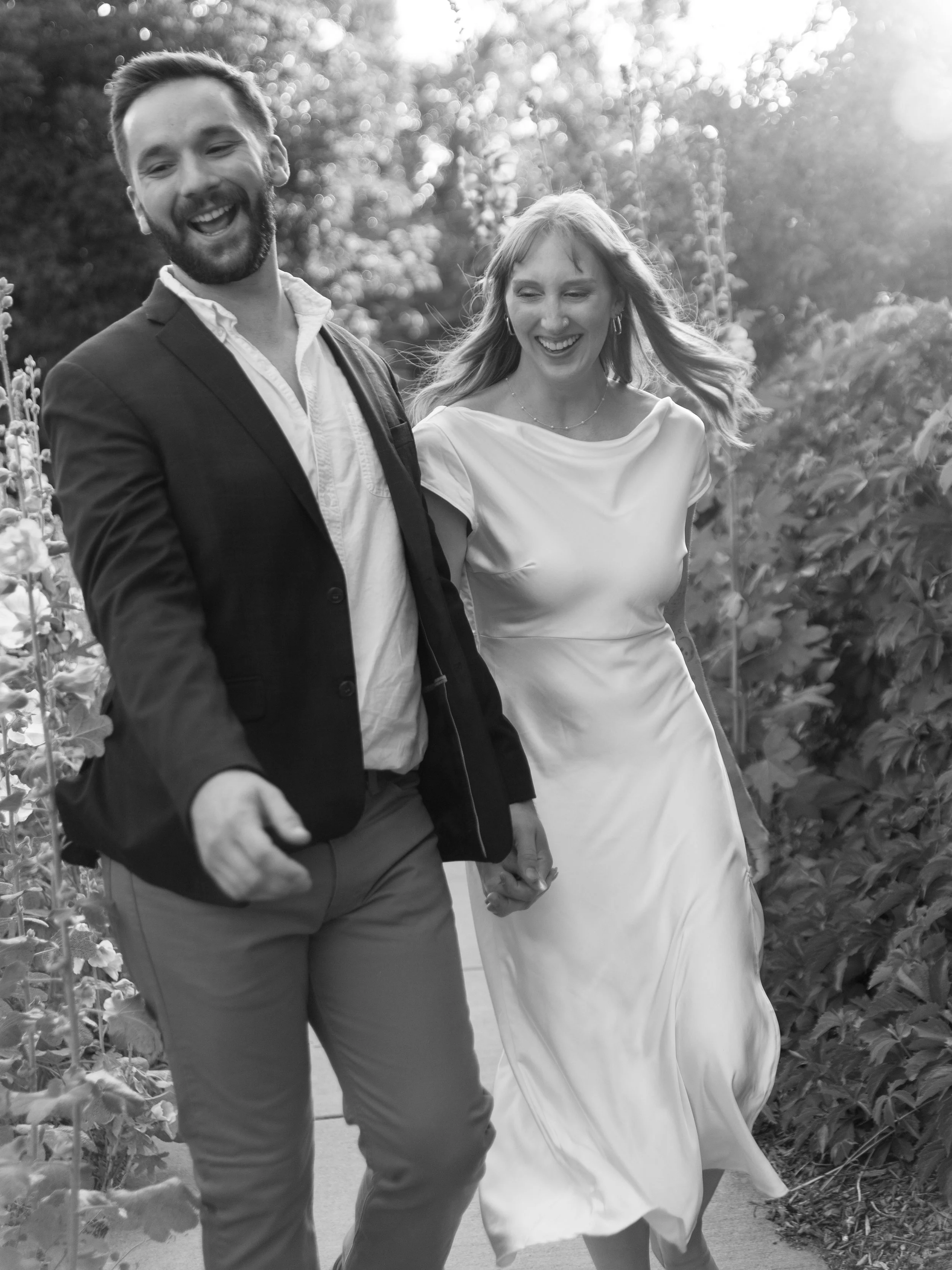 A smiling couple holding hands and walking outdoors. The man is wearing a dark blazer and light-colored pants, and the woman is in a flowing white dress. They are surrounded by trees and sunlight.