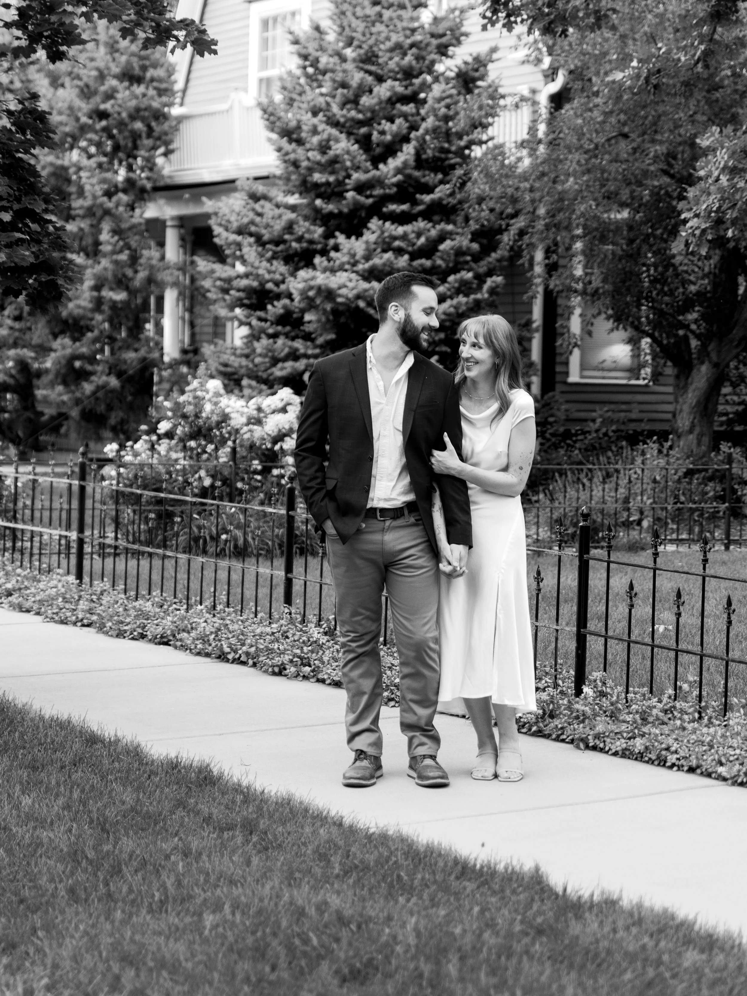 A couple walking hand-in-hand along a sidewalk, smiling at each other, with trees and a house in the background.