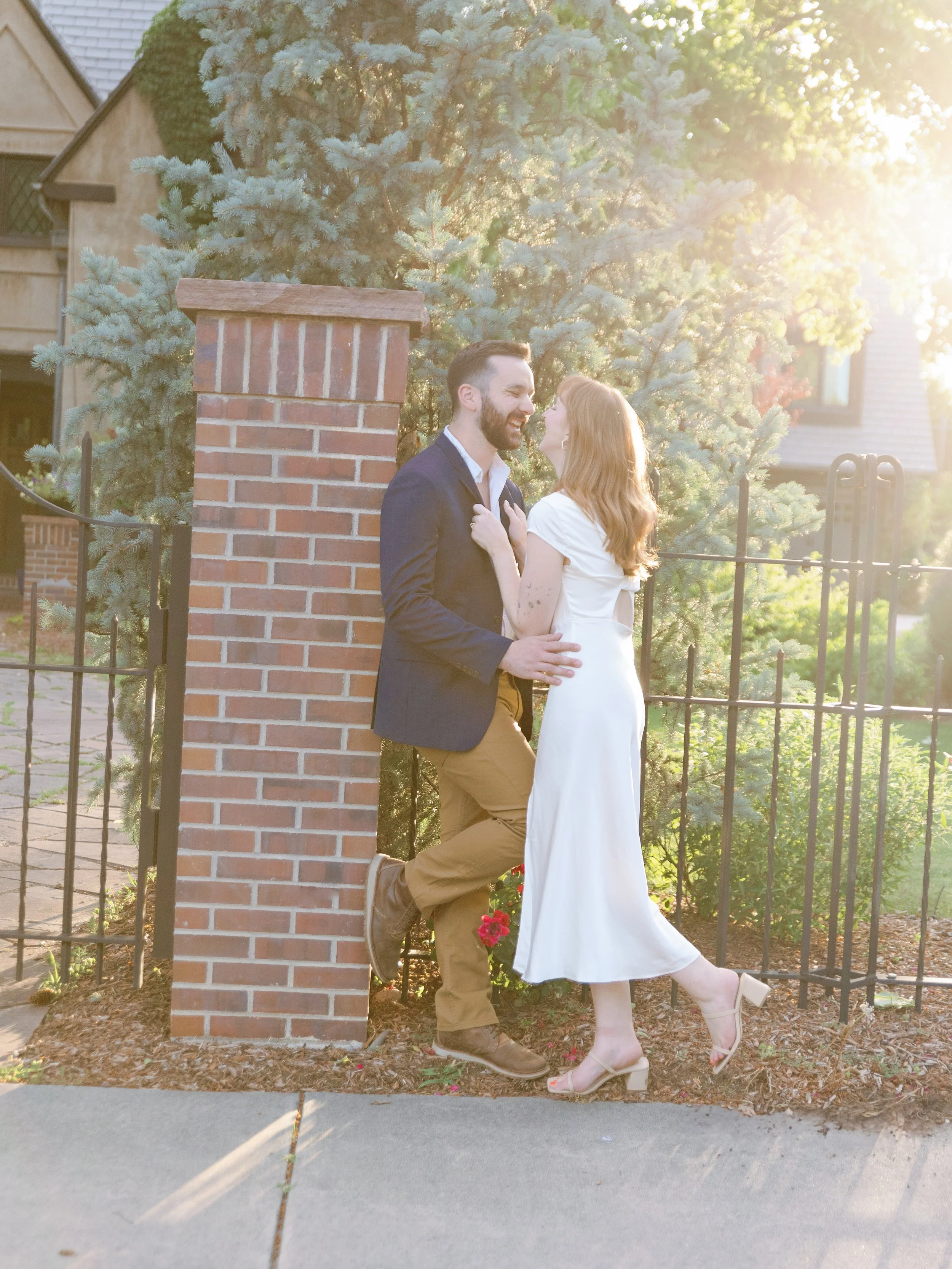 A couple standing and leaning on a brick pillar, smiling and looking at each other, in front of a tall pine tree and a metal fence at sunset.