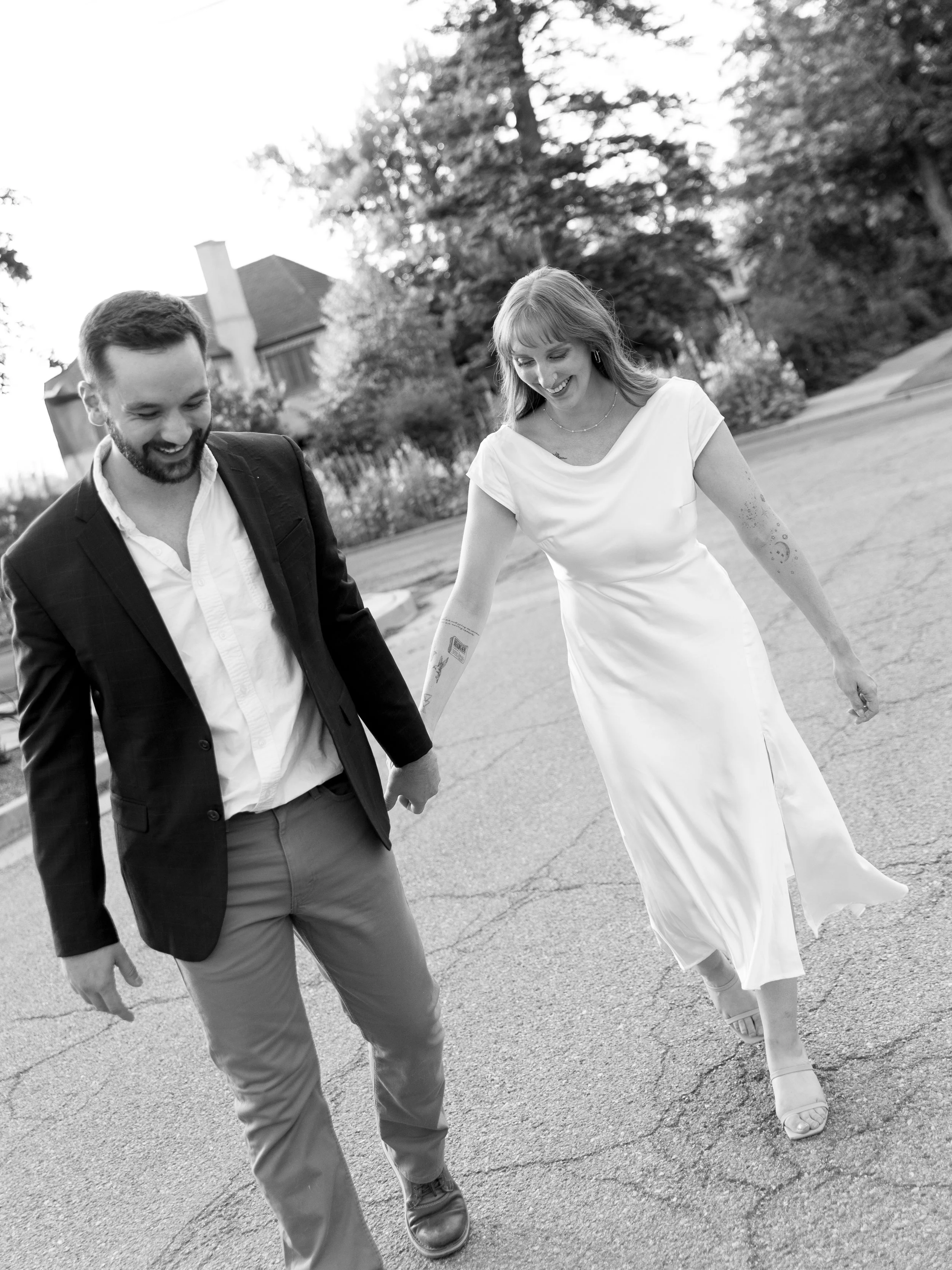 A man and woman walking hand in hand outdoors, smiling and looking down, with trees and houses in the background.