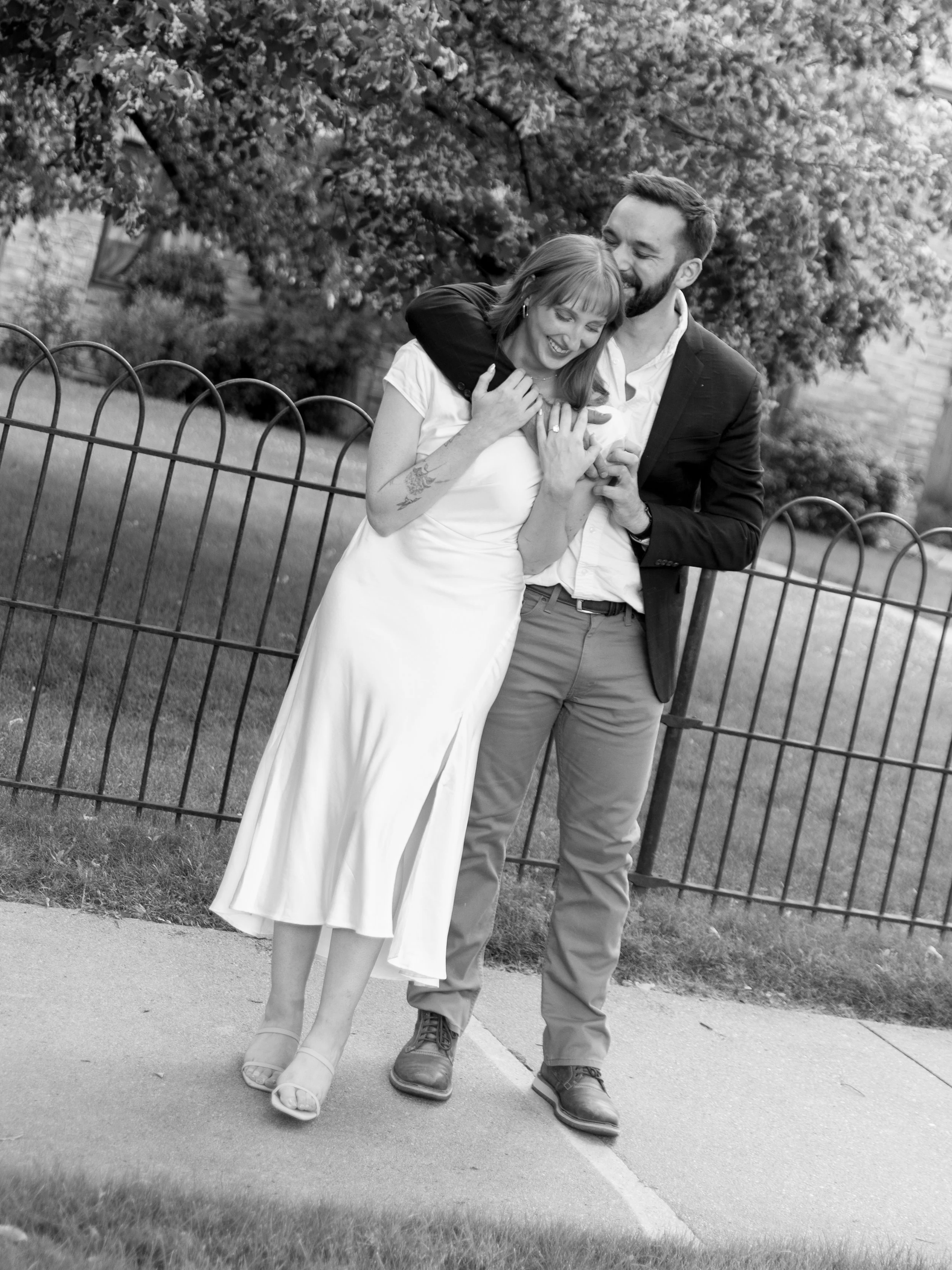 A couple, the woman in a white dress and the man in a blazer and trousers, embracing and laughing outside near a fence and trees.