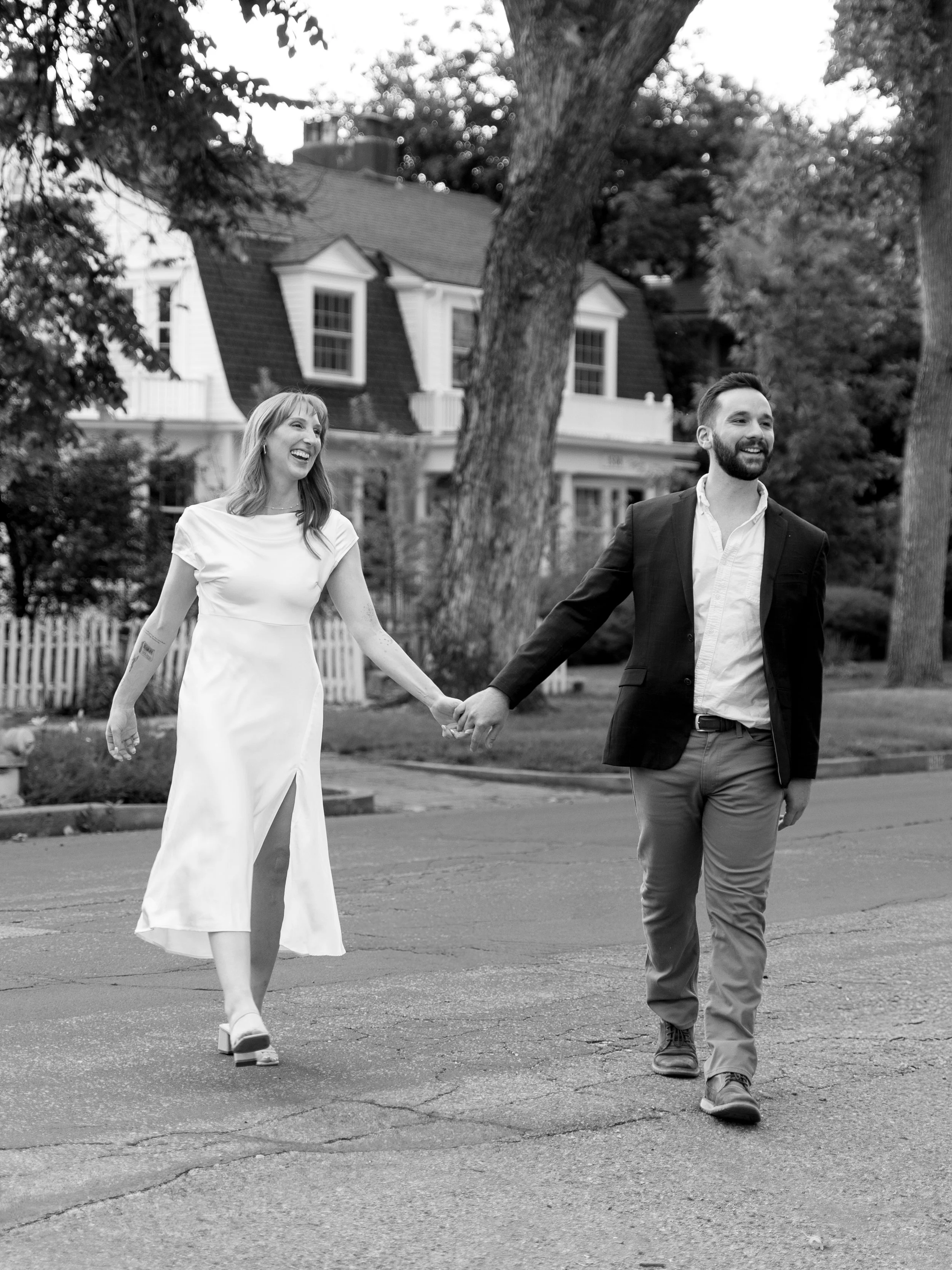 A smiling woman in a white dress and a man in a suit walking hand in hand on a street, with trees and a house in the background.