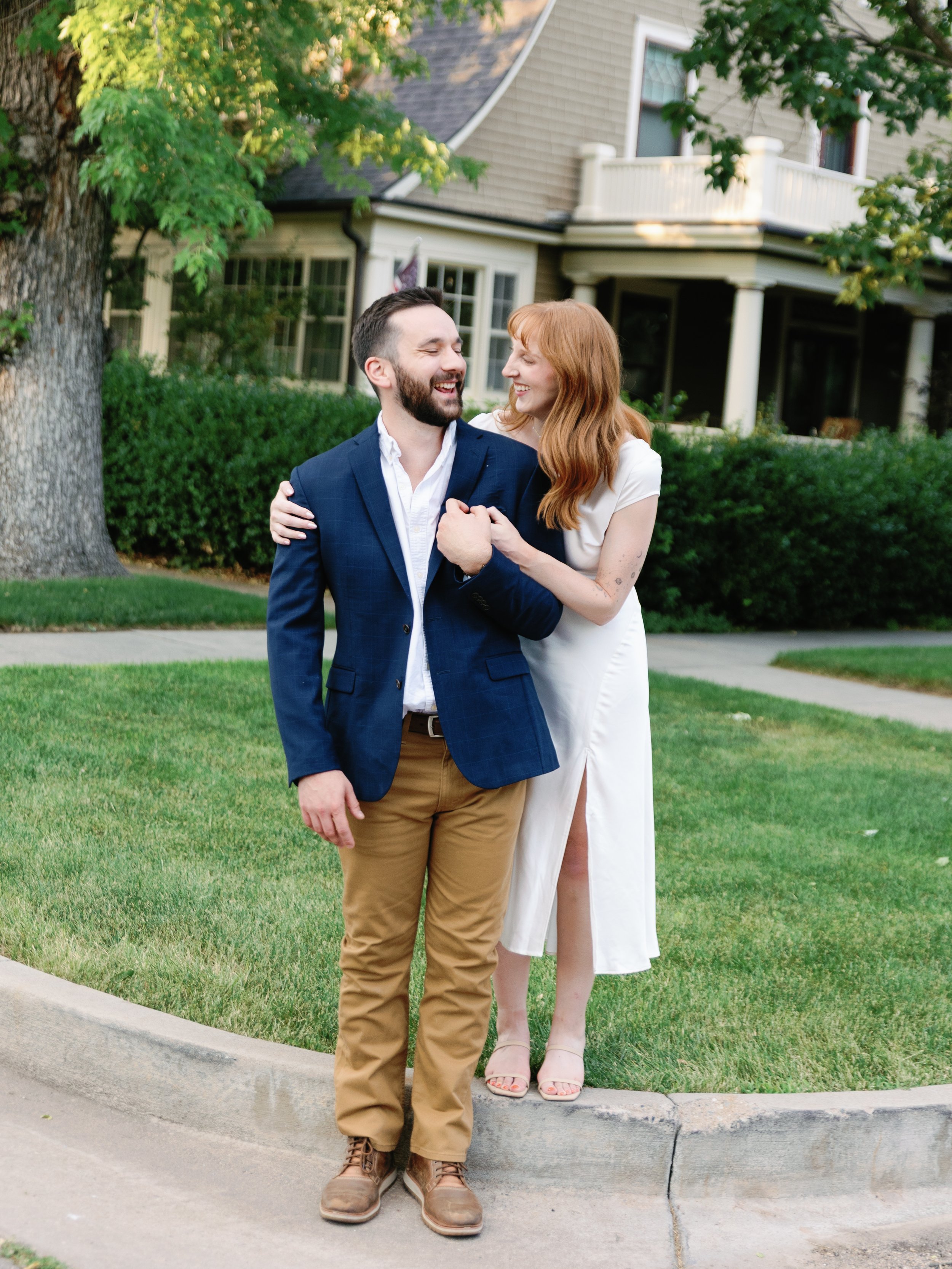 A young couple is standing on a sidewalk in front of a house, smiling and laughing, with greenery and a tree in the background. The man is wearing a blue blazer, white shirt, and khaki pants. The woman is wearing a white dress with a slit and heels.