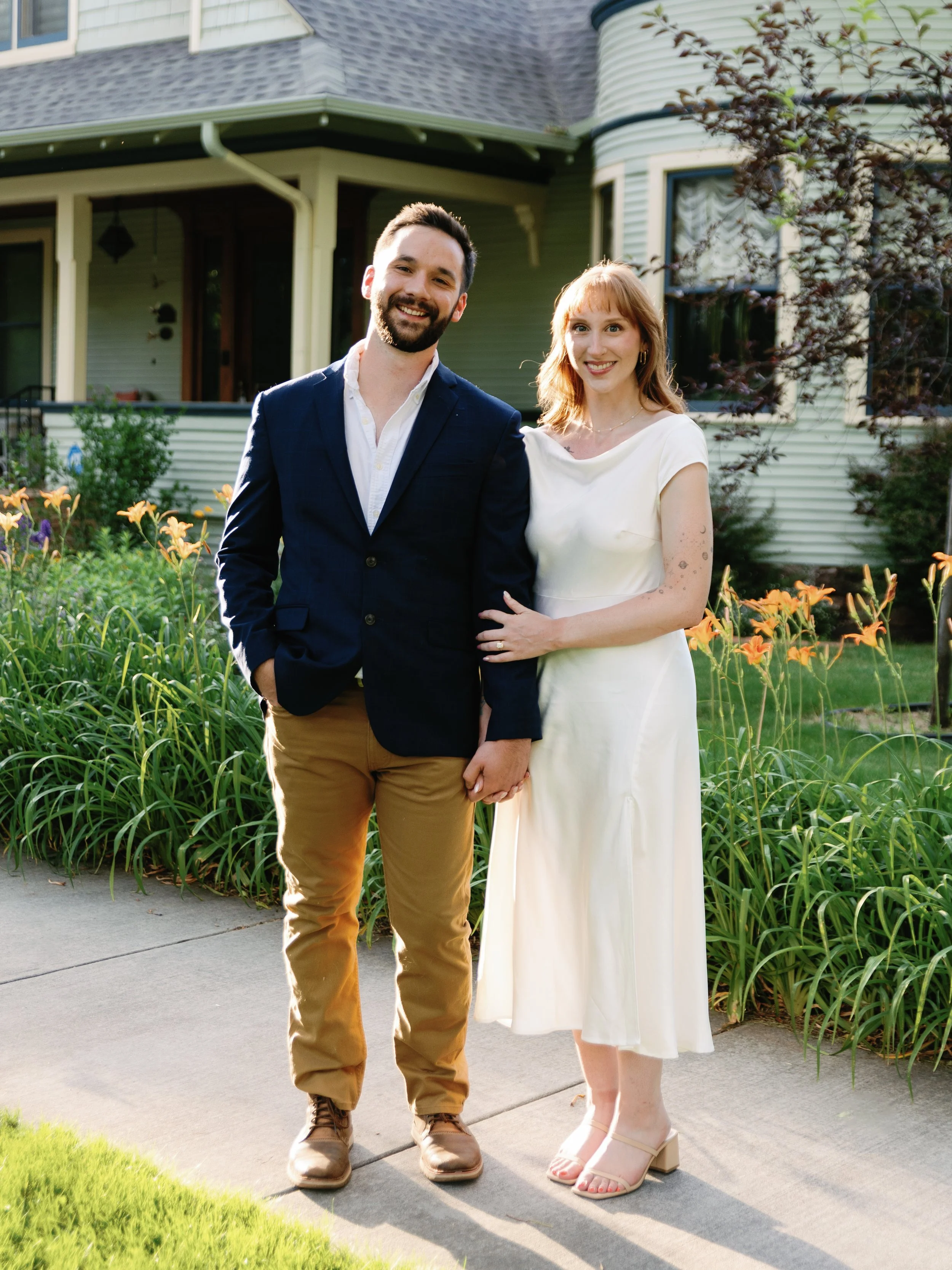 A smiling man and woman holding hands, standing on a sidewalk in front of a house with a garden, during daylight.