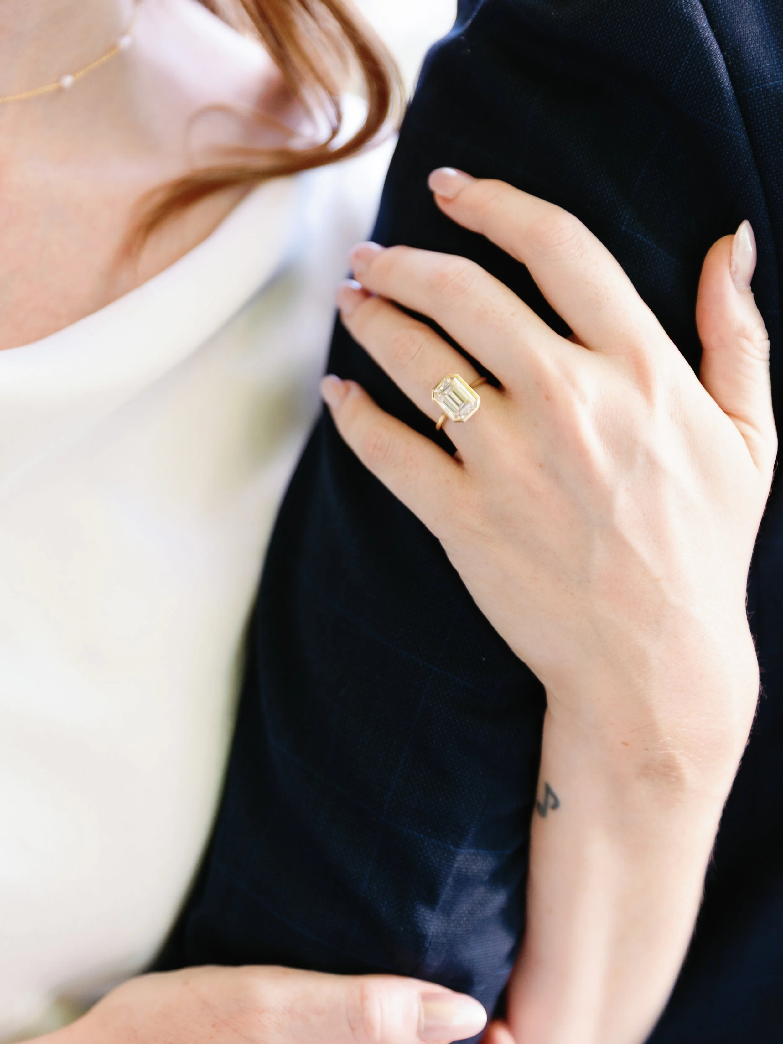 A woman with a gold ring featuring a large, rectangular emerald-cut gemstone on her finger, rests her hand on a man's shoulder.