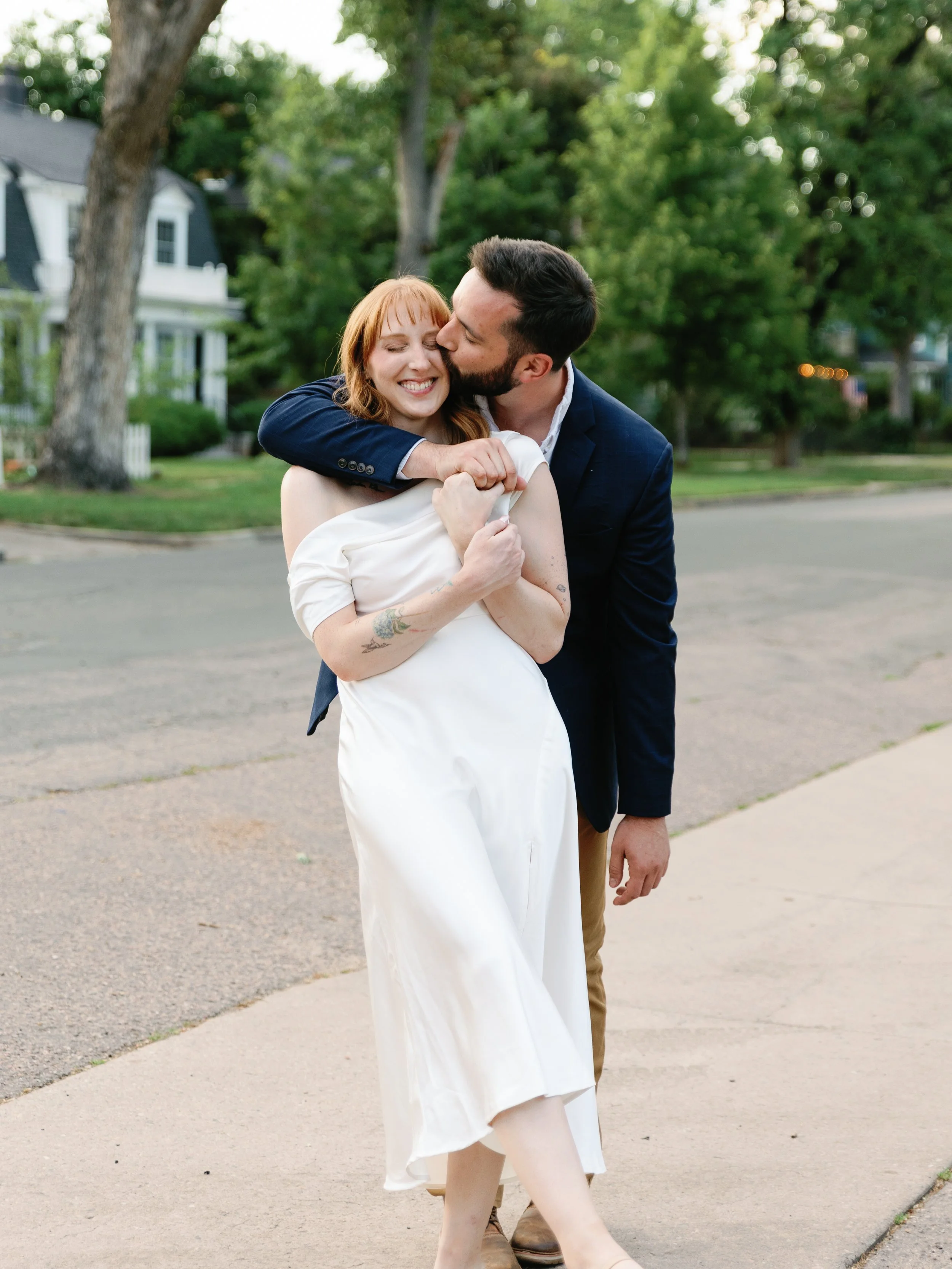 A couple hugging on a sidewalk in a residential neighborhood, with trees and houses in the background. The woman is wearing a white dress and the man is dressed in a navy blazer and khakis. The man is kissing the woman's cheek while she smiles with e