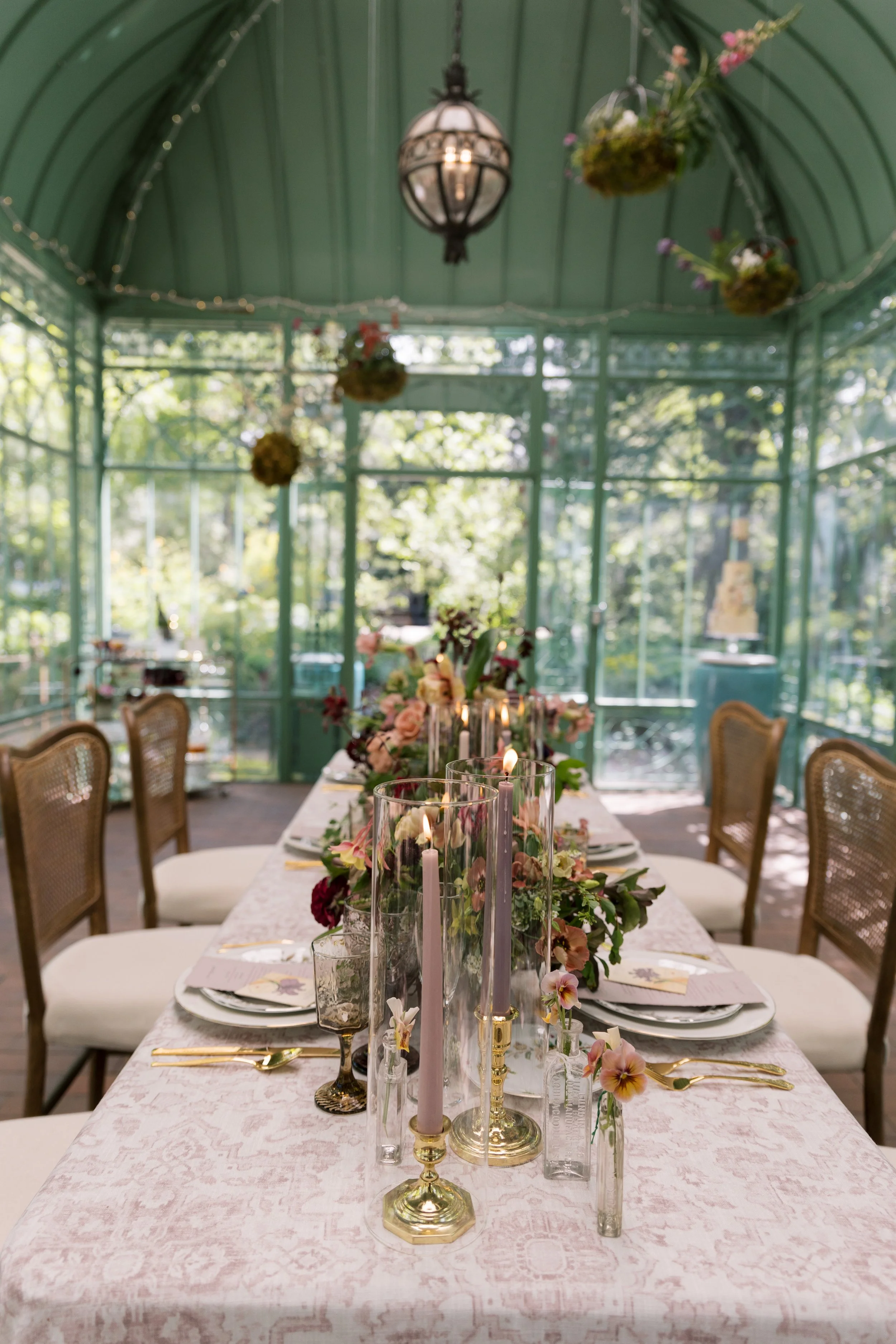 A long dining table decorated with candles, flowers, and elegant tableware inside a glass greenhouse with greenery outside.