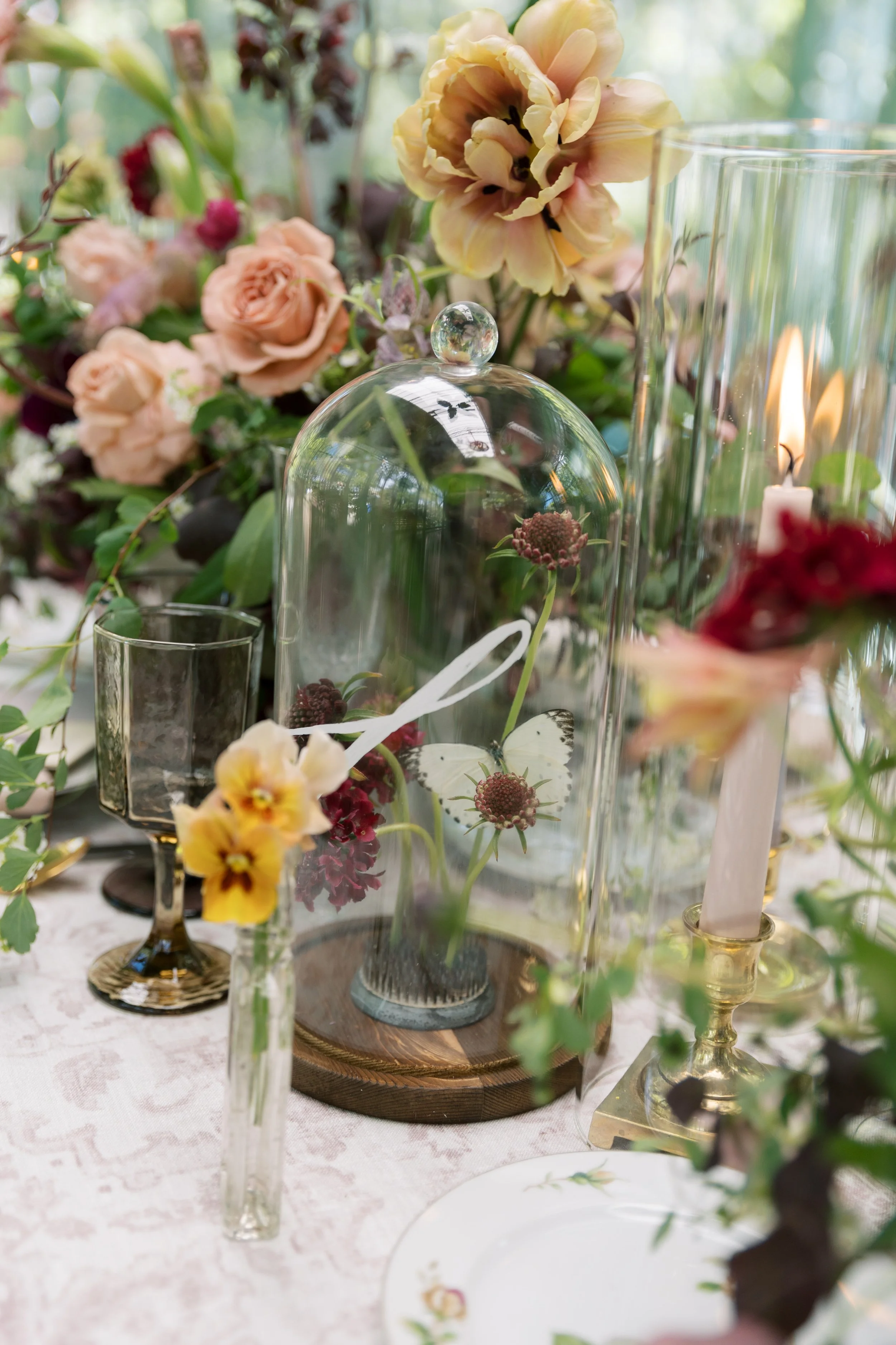 A table centerpiece with a glass dome covering flowers, surrounded by candles, a vase with yellow and pink flowers, and a floral arrangement with peonies and roses.