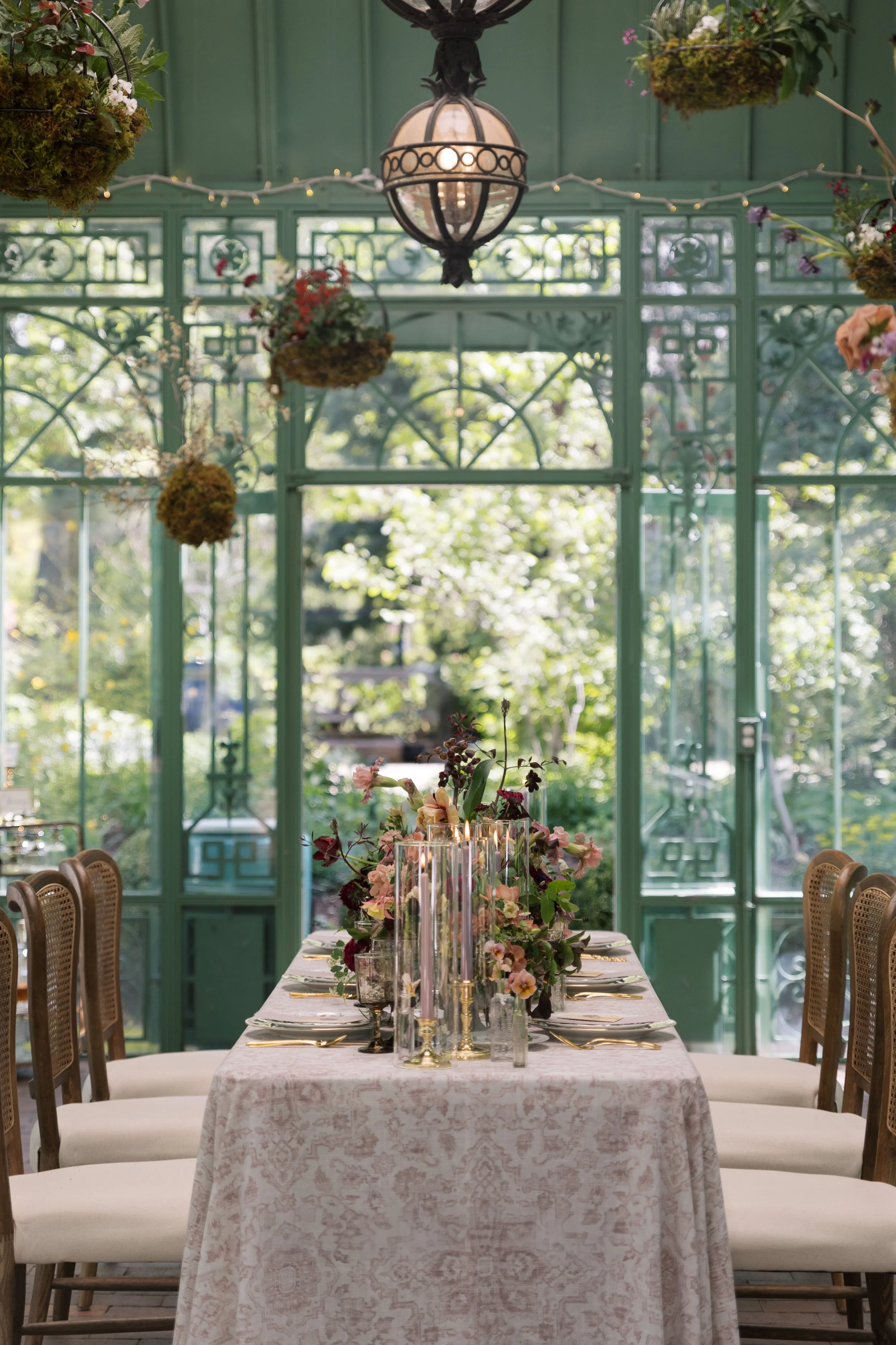 A dining table set with a floral centerpiece, surrounded by chairs, inside a green enclosed patio with hanging plants and a vintage chandelier.
