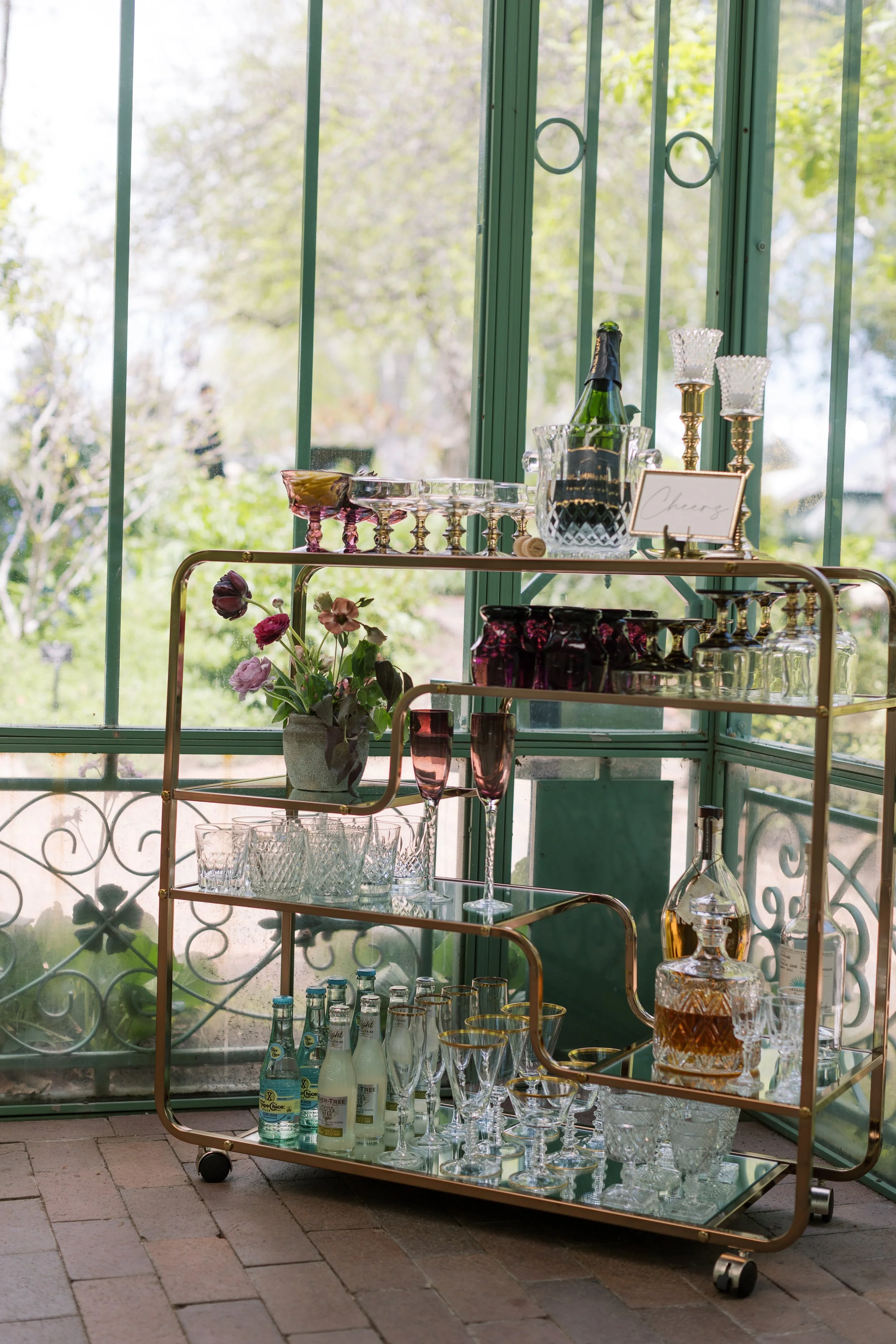 A gold bar cart with glassware, bottles, and a flower arrangement near a large window with green frames, overlooking trees and a garden.