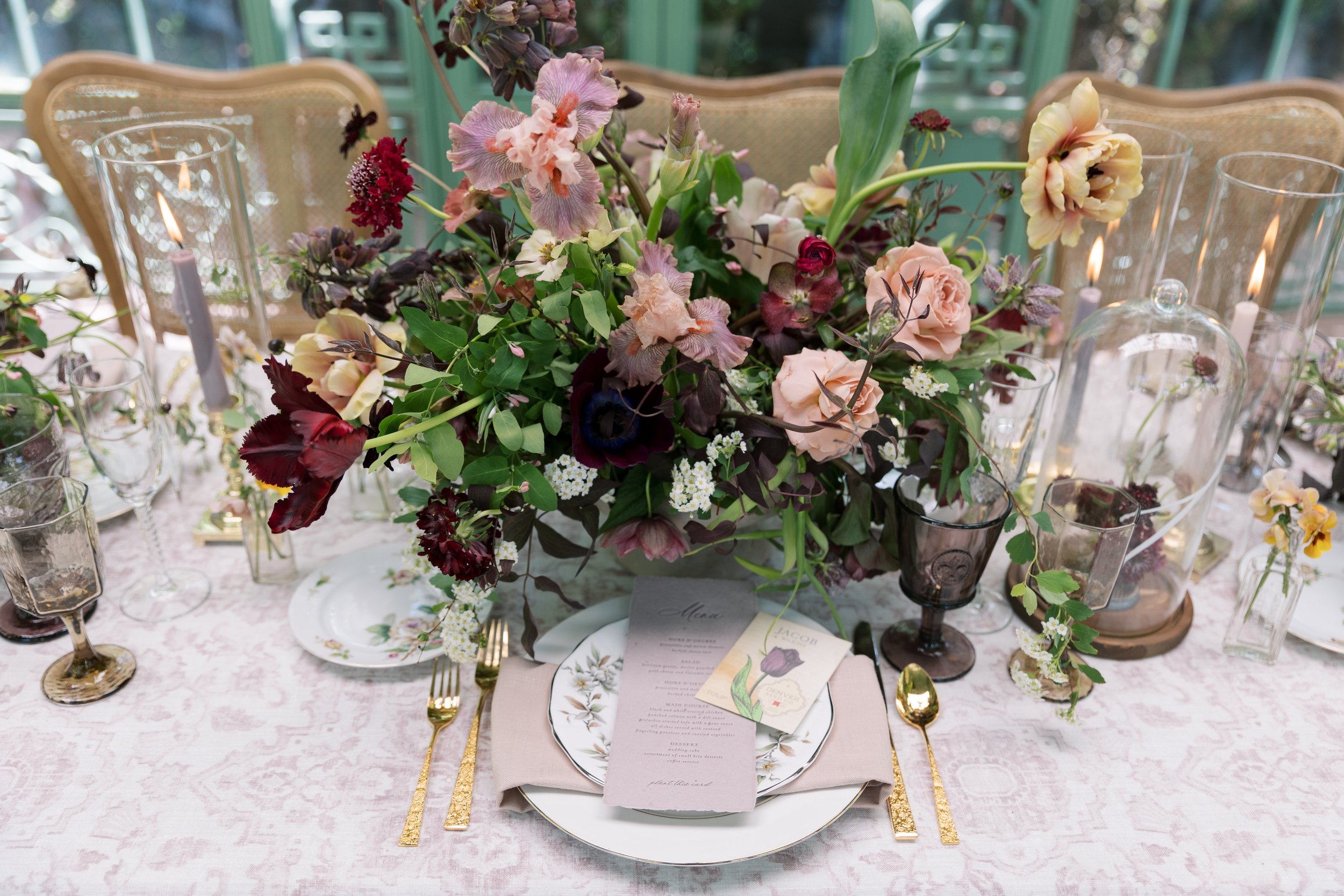 A table setting with a floral centerpiece, gold utensils, and decorative candles in glass holders on a pink tablecloth.