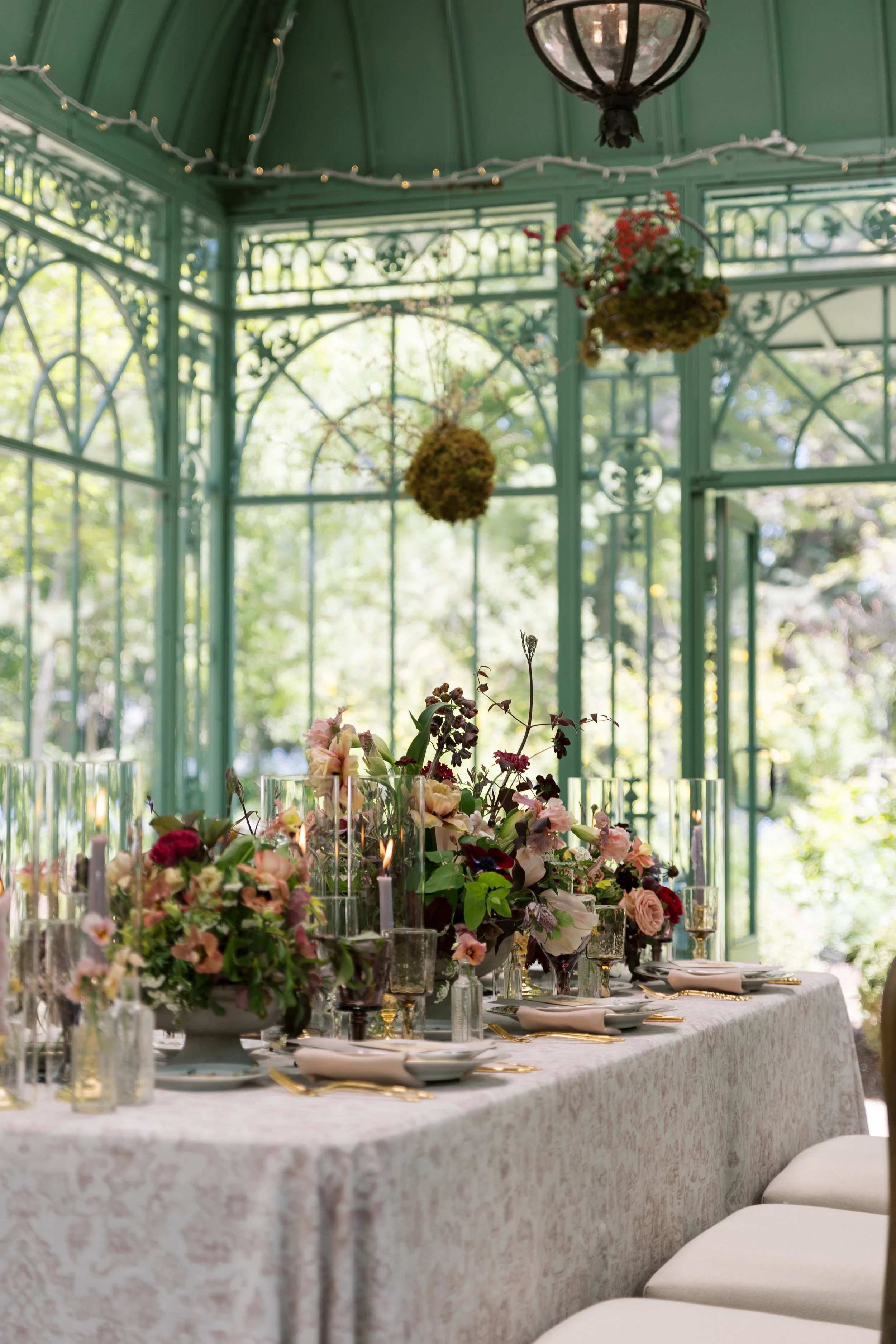 A elegantly decorated banquet table with floral centerpieces, candles, and tableware in a glass conservatory filled with natural light and hanging plants.