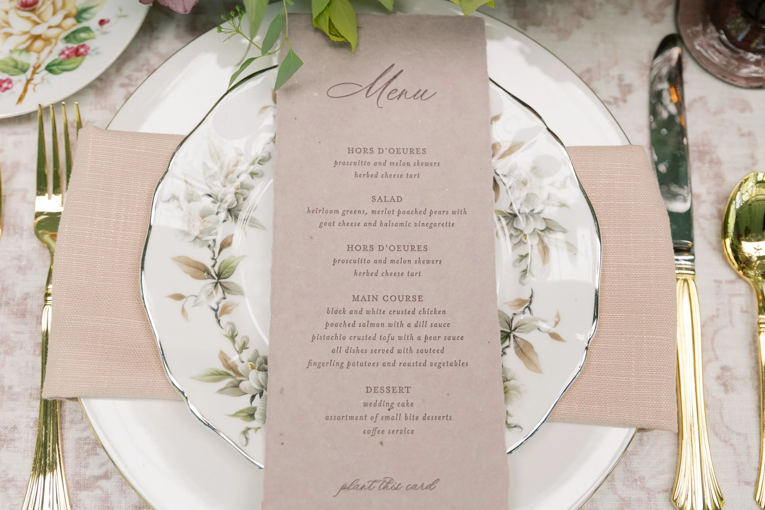 Elegant place setting with a floral-patterned porcelain plate, a pink napkin, gold cutlery, and a menu card on a pink tablecloth, with greenery and a glass in the background.