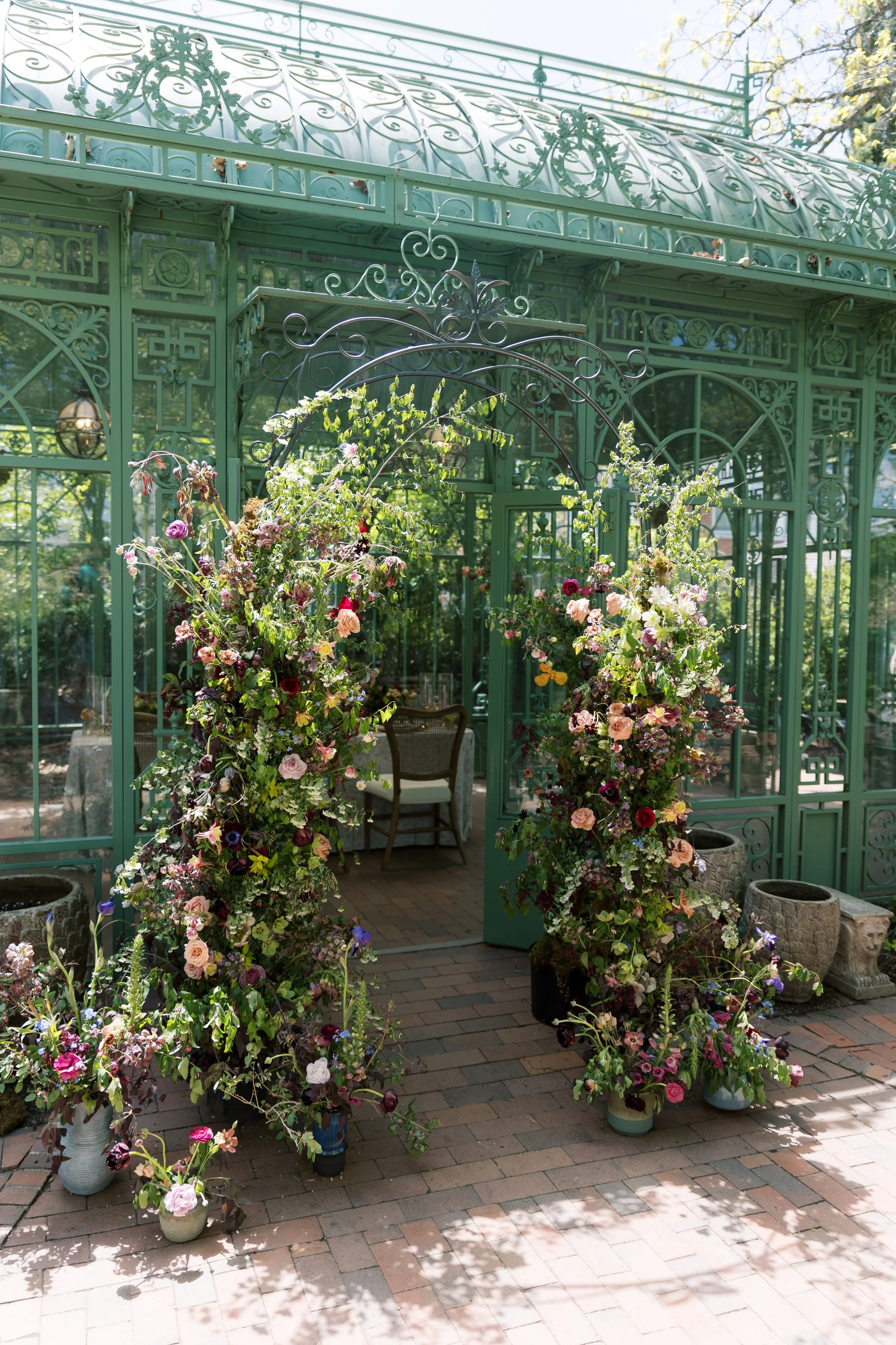 Green ornate glass greenhouse with two flower arrangements on either side of an arched entrance, inside a garden setting with potted plants on the brick ground.
