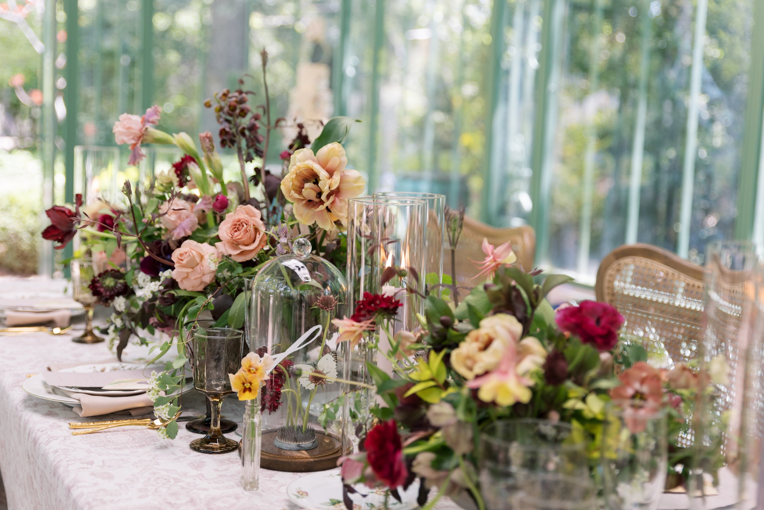 A beautifully set dining table with floral centerpieces, glass dome, and elegant tableware, in a bright, sunlit room surrounded by large windows.