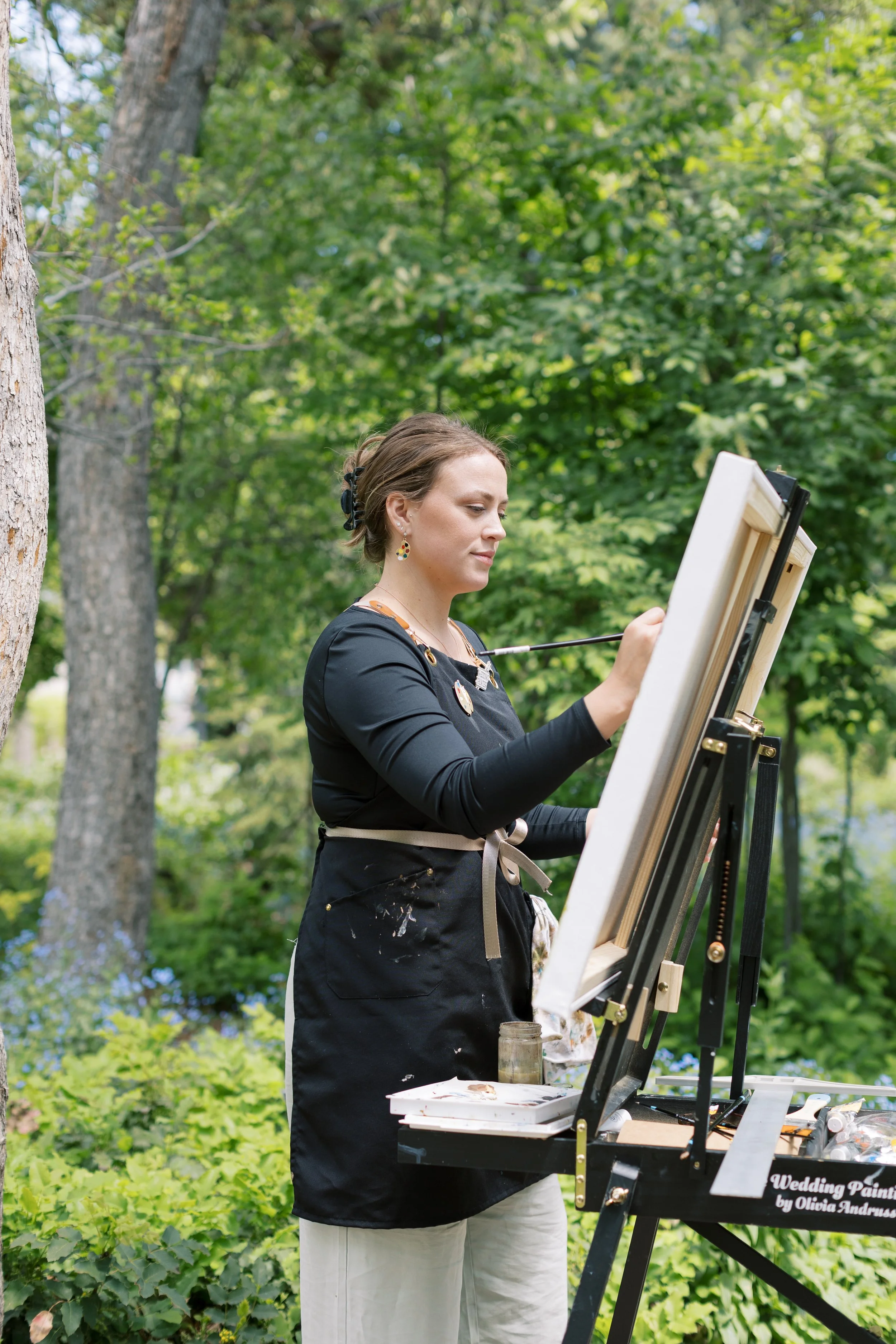 A woman painting outdoors on a canvas set up on an easel, surrounded by green trees and foliage.