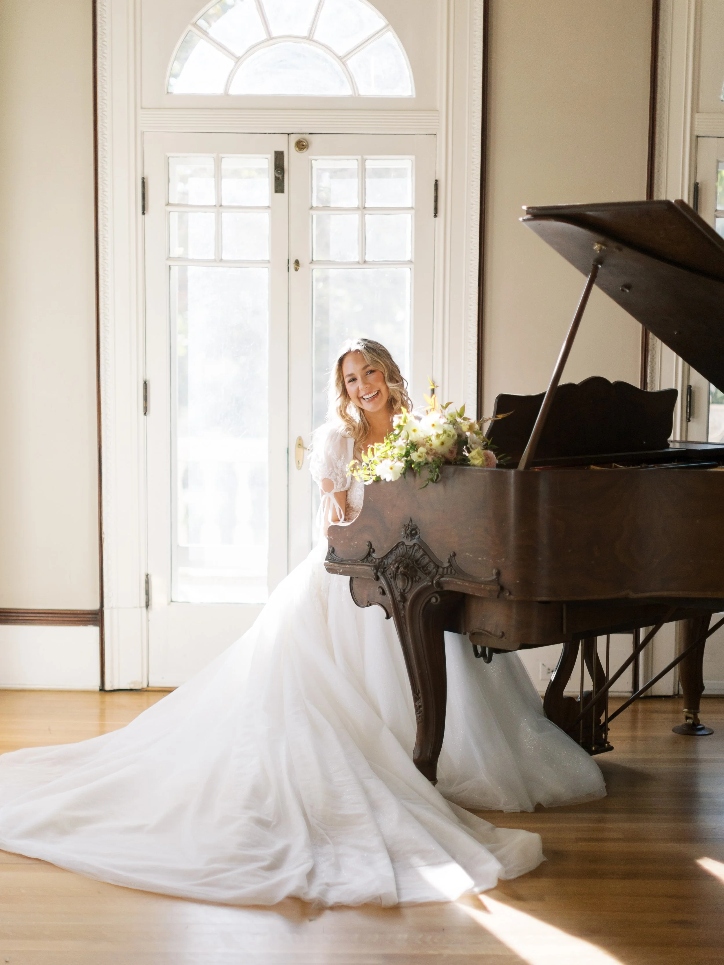 A bride in a white wedding gown smiling and leaning on a grand piano with a floral arrangement, in front of a large window.