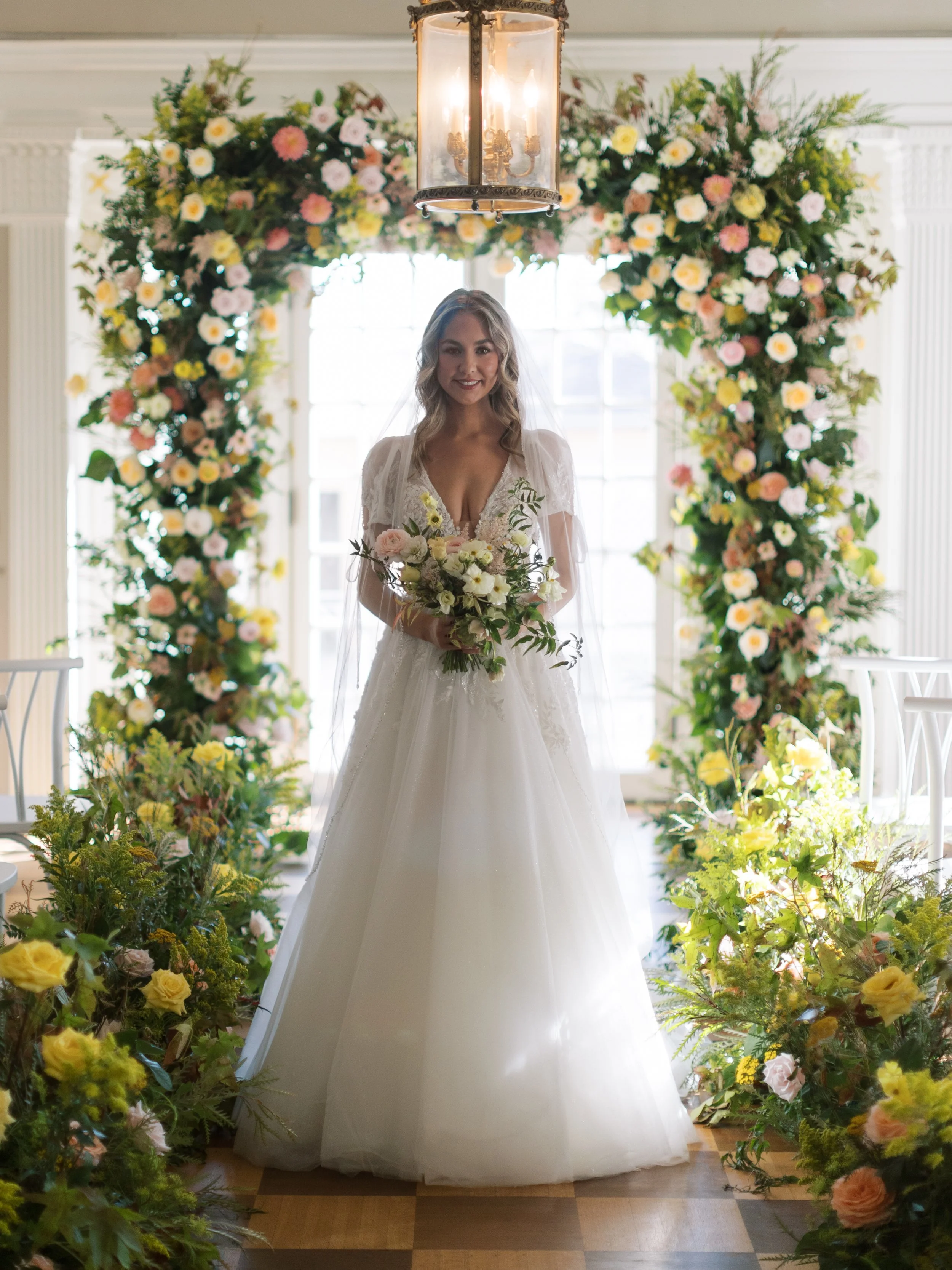 A bride standing under a floral archway in a bright room, holding a bouquet of flowers, with sunlight streaming through large windows in the background.