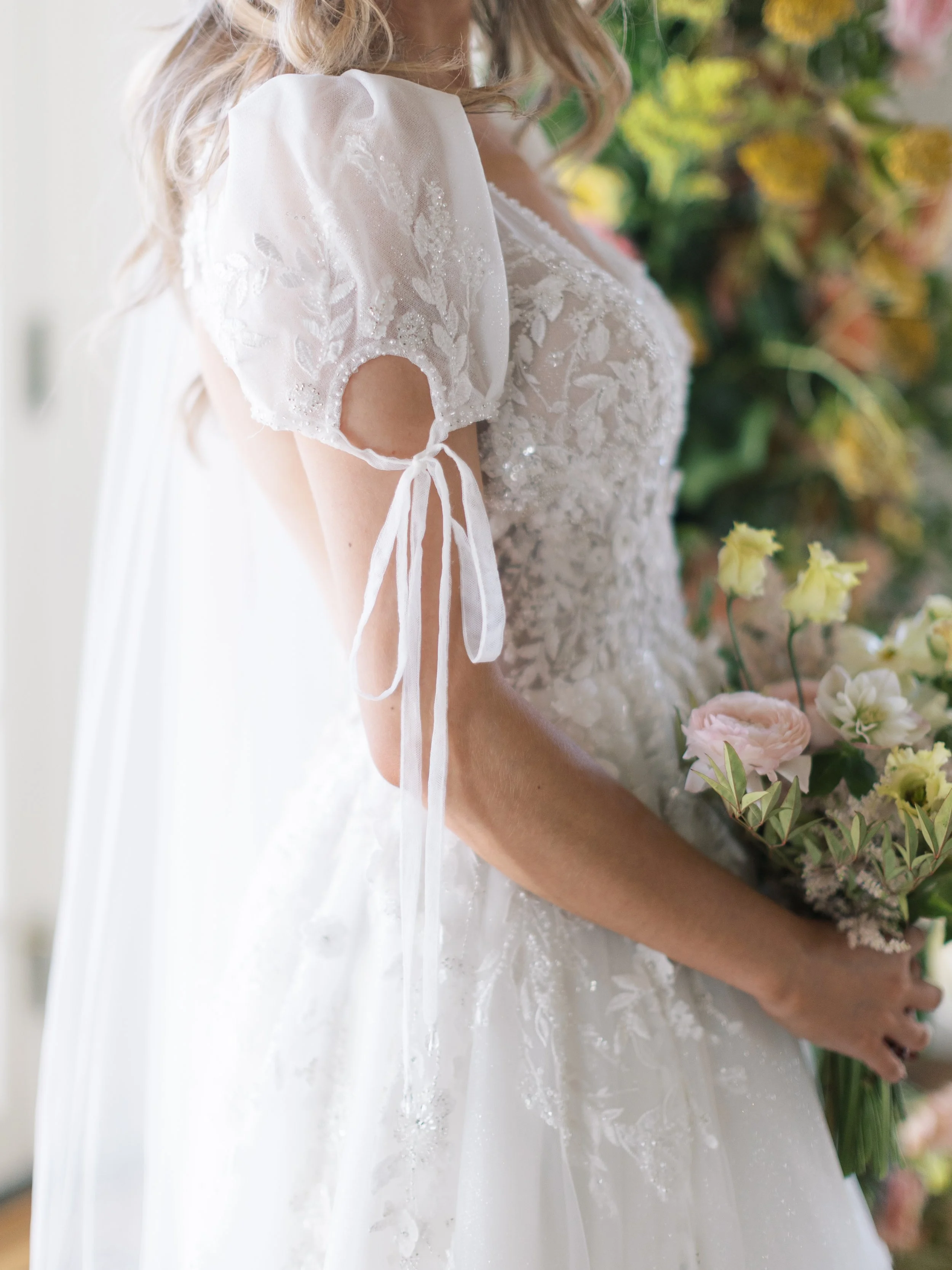 Close-up of a bride holding a bouquet of flowers, wearing a white wedding dress with lace embroidery and puffed sleeves with ribbon ties.
