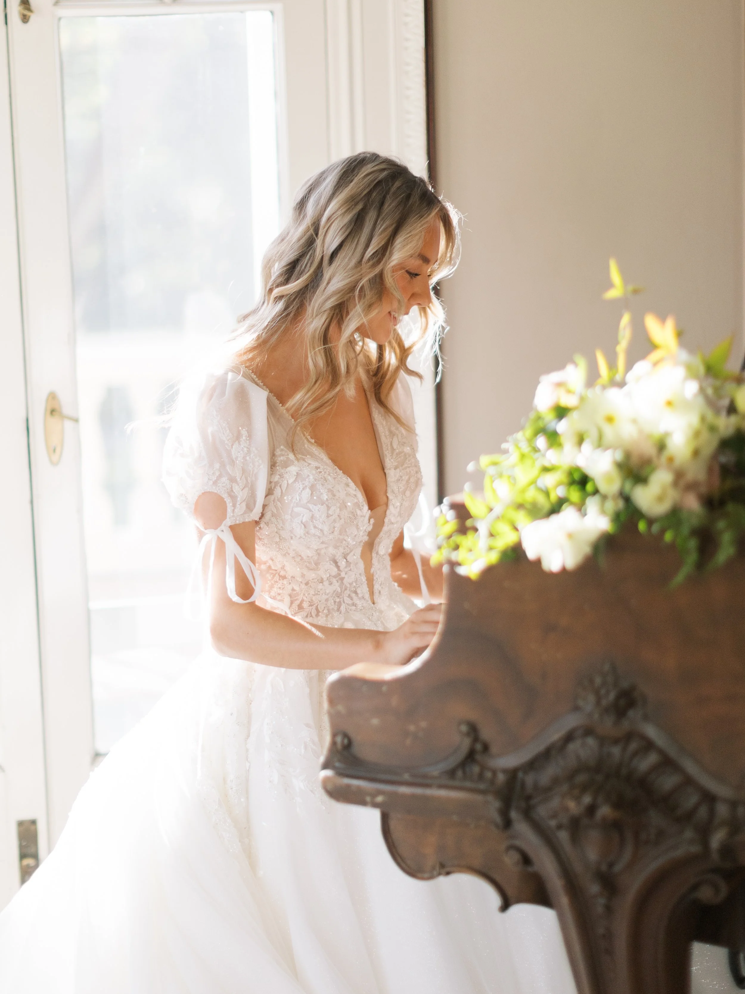 A bride with blonde, wavy hair in a white lace wedding dress, playing the piano in a sunlit room near a glass door with a bouquet of white and yellow flowers on the piano.