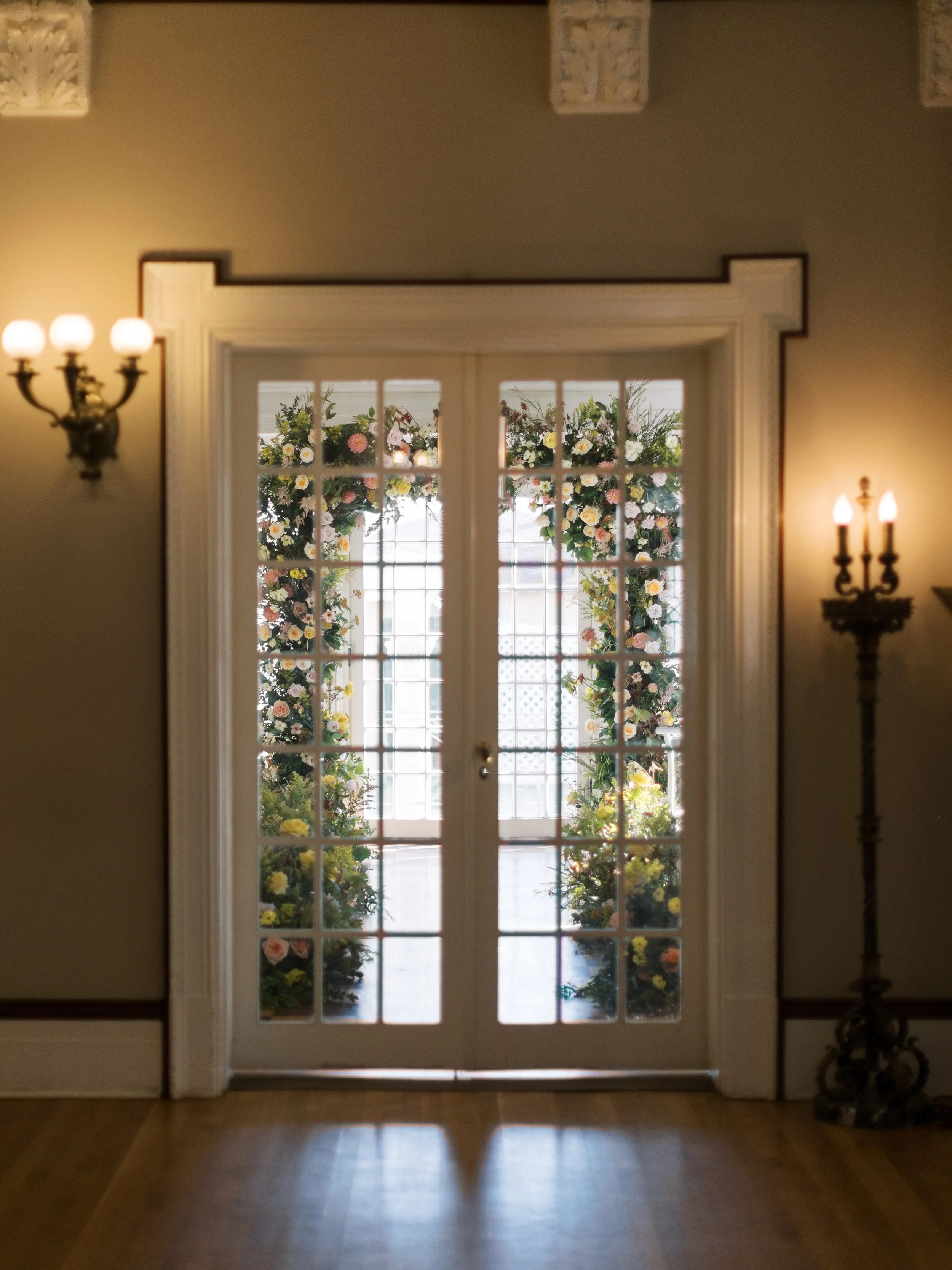 Double glass door with flowers on the outside, flanked by wall sconces with lit bulbs inside.