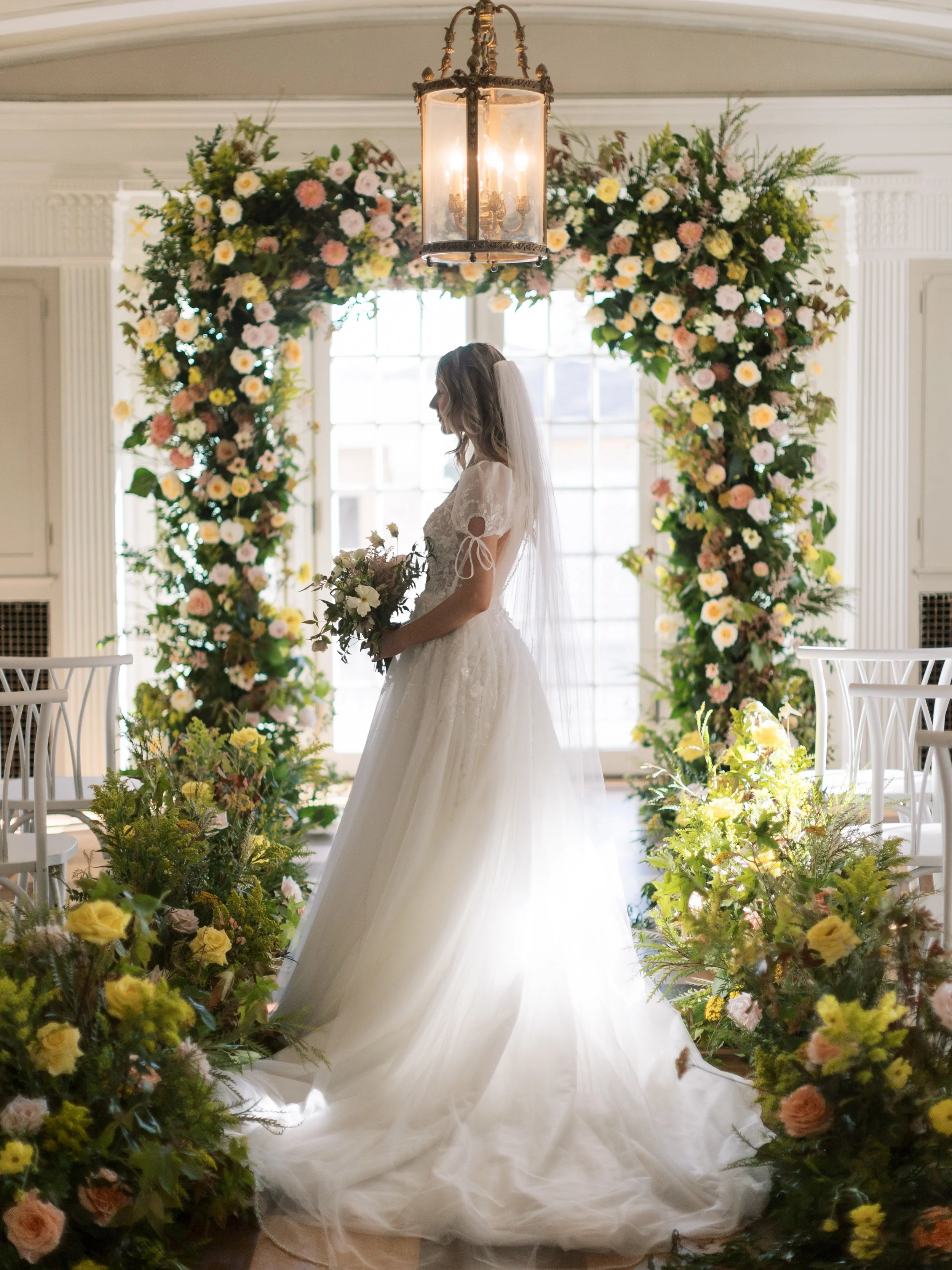 A bride in a white wedding gown holding a bouquet, standing under a floral arch with a chandelier, in a bright room with large windows and white chairs.