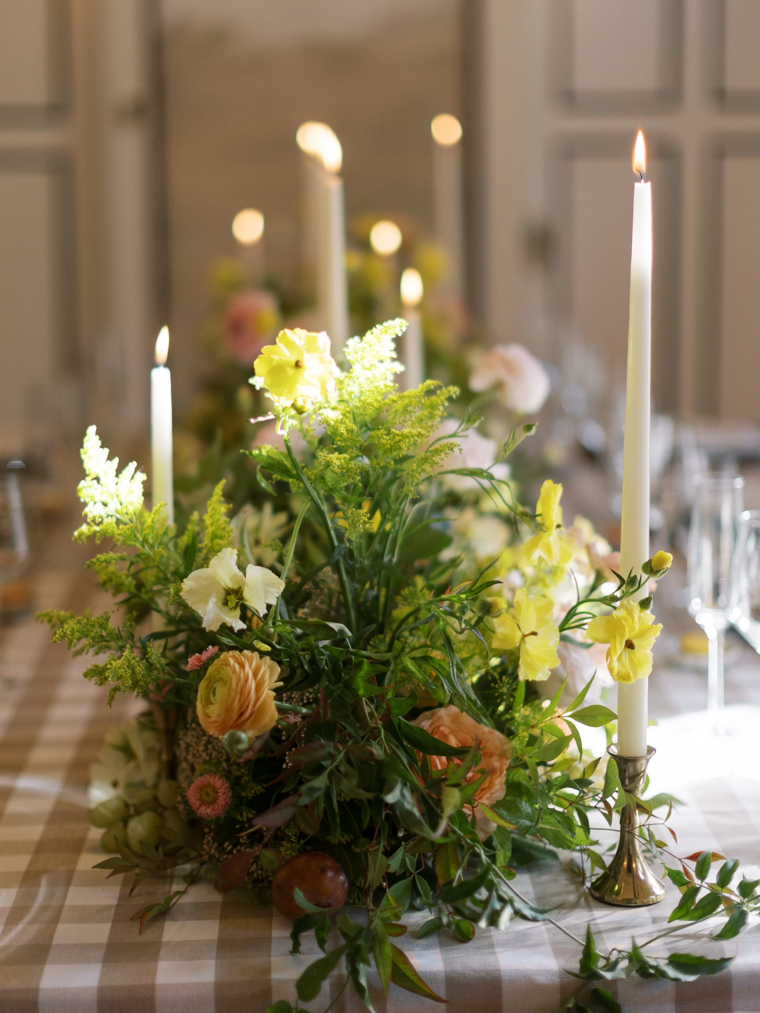 Elegant table centerpiece with yellow and white flowers, greenery, and lit tall white candles on a checkered tablecloth.