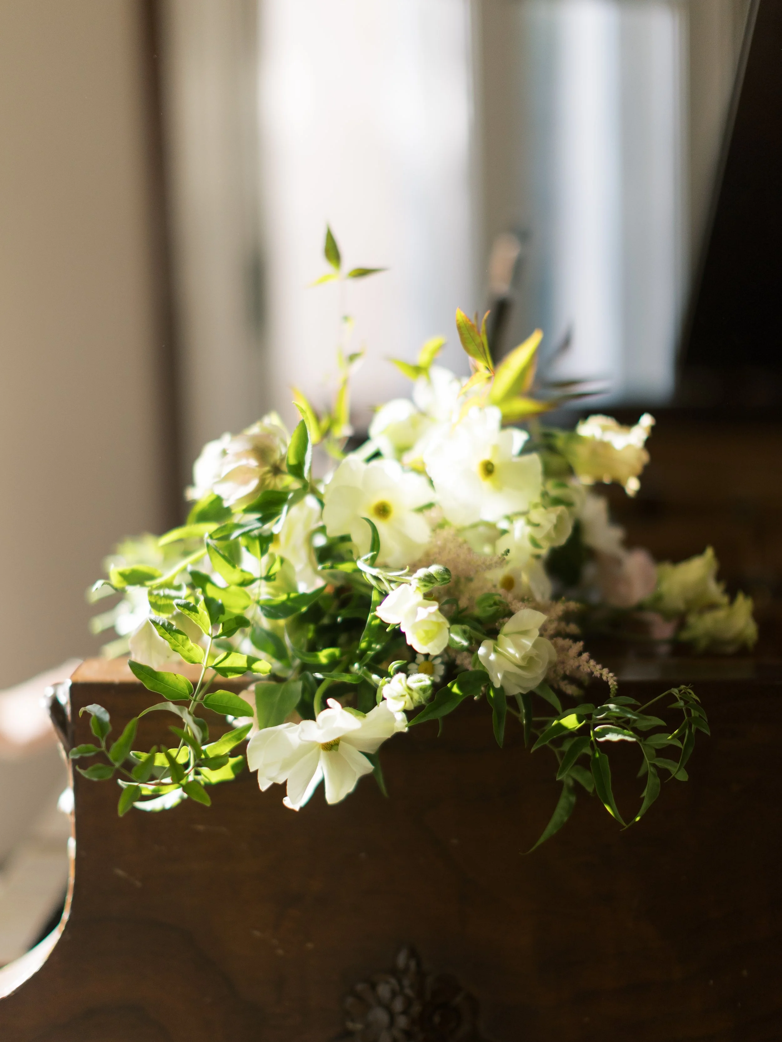 A bouquet of white and pale yellow flowers with green leaves on a wooden surface near a window with bright sunlight.
