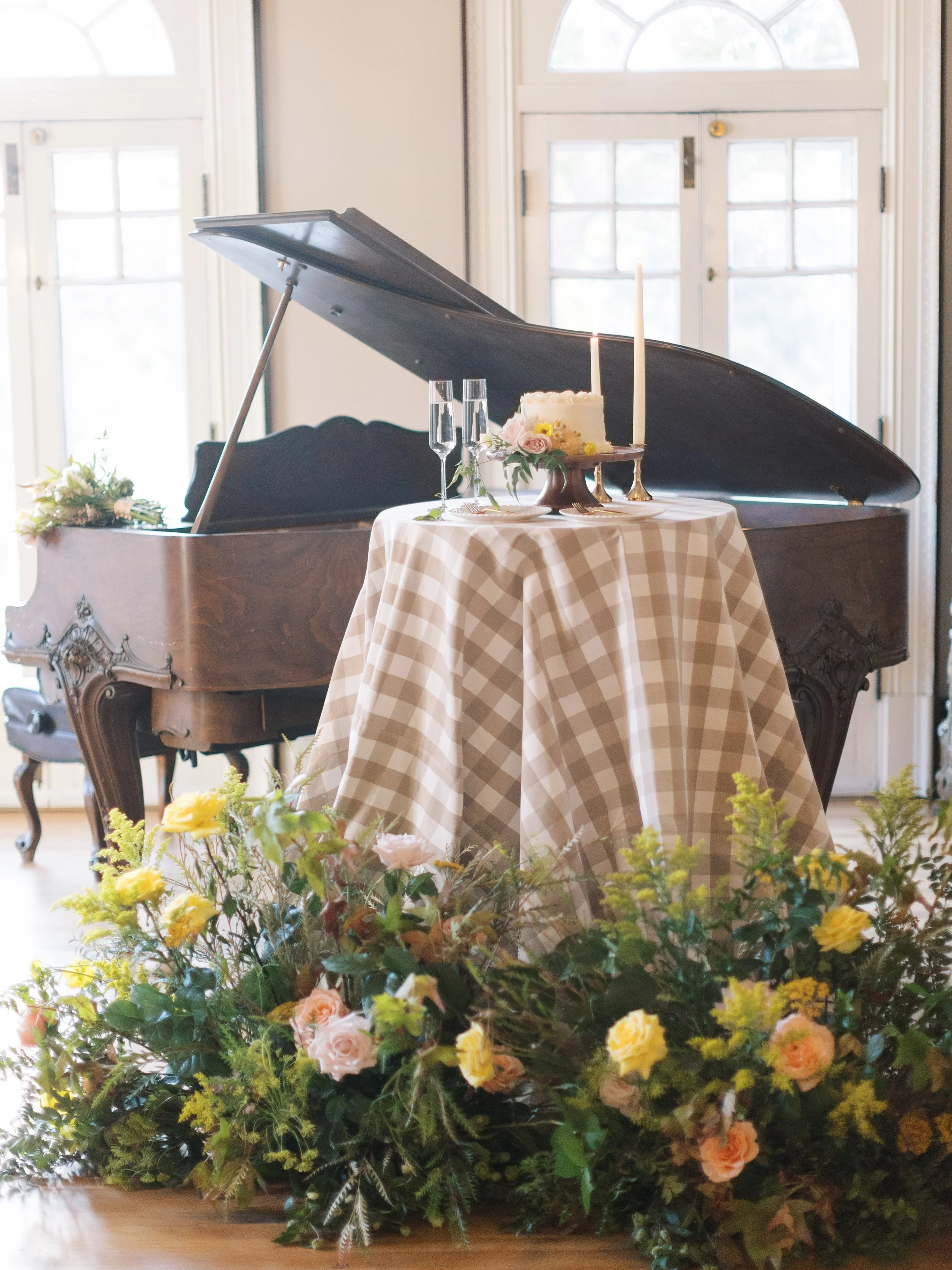 A grand piano in a bright room with large windows, a small round table with a checked tablecloth, a decorated cake with lit candles, champagne glasses, and a floral arrangement in front.