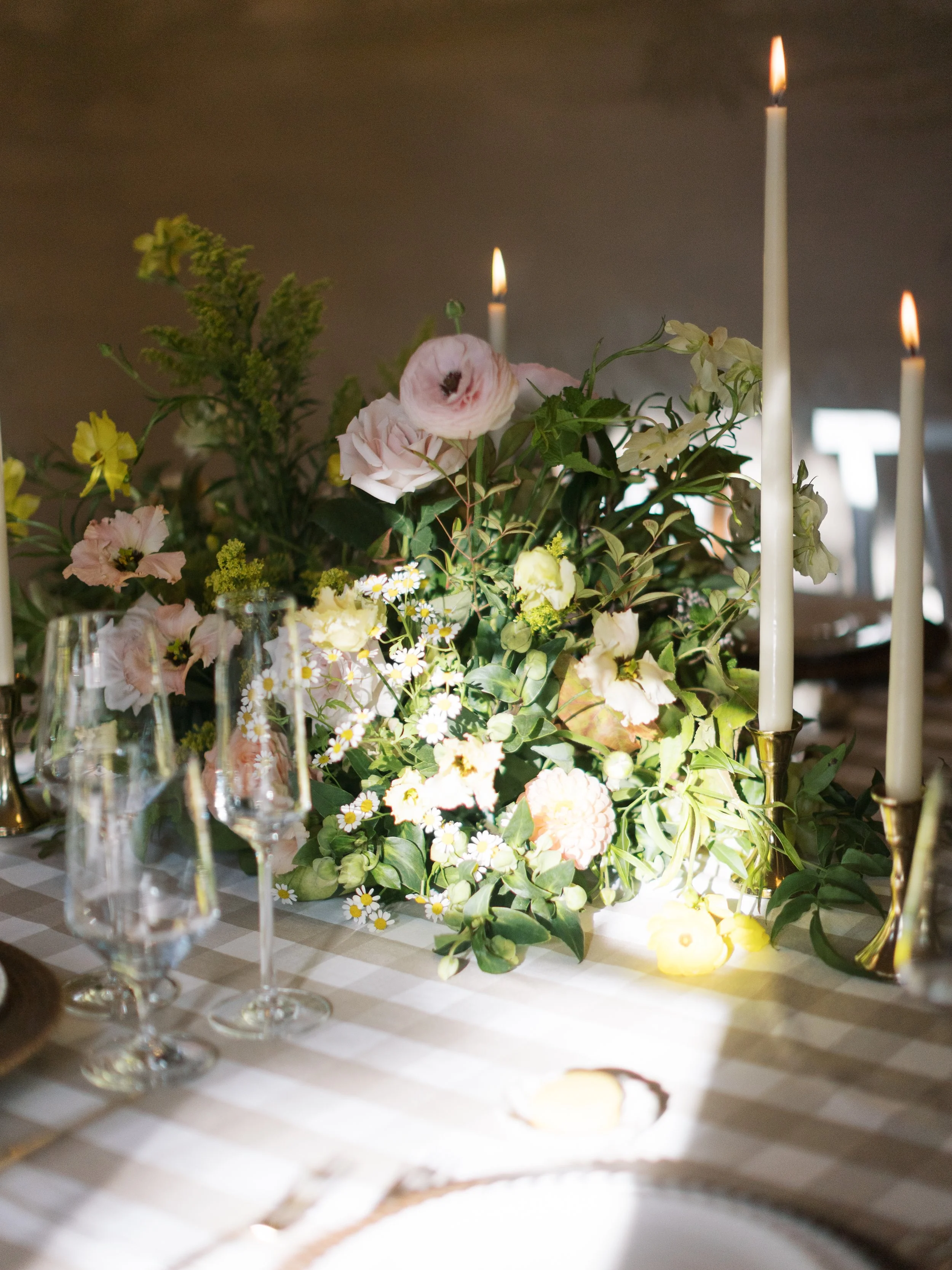 A table decorated with a floral centerpiece of pink, yellow, and white flowers, several lit white candles in gold candleholders, and glassware on a checkered tablecloth.
