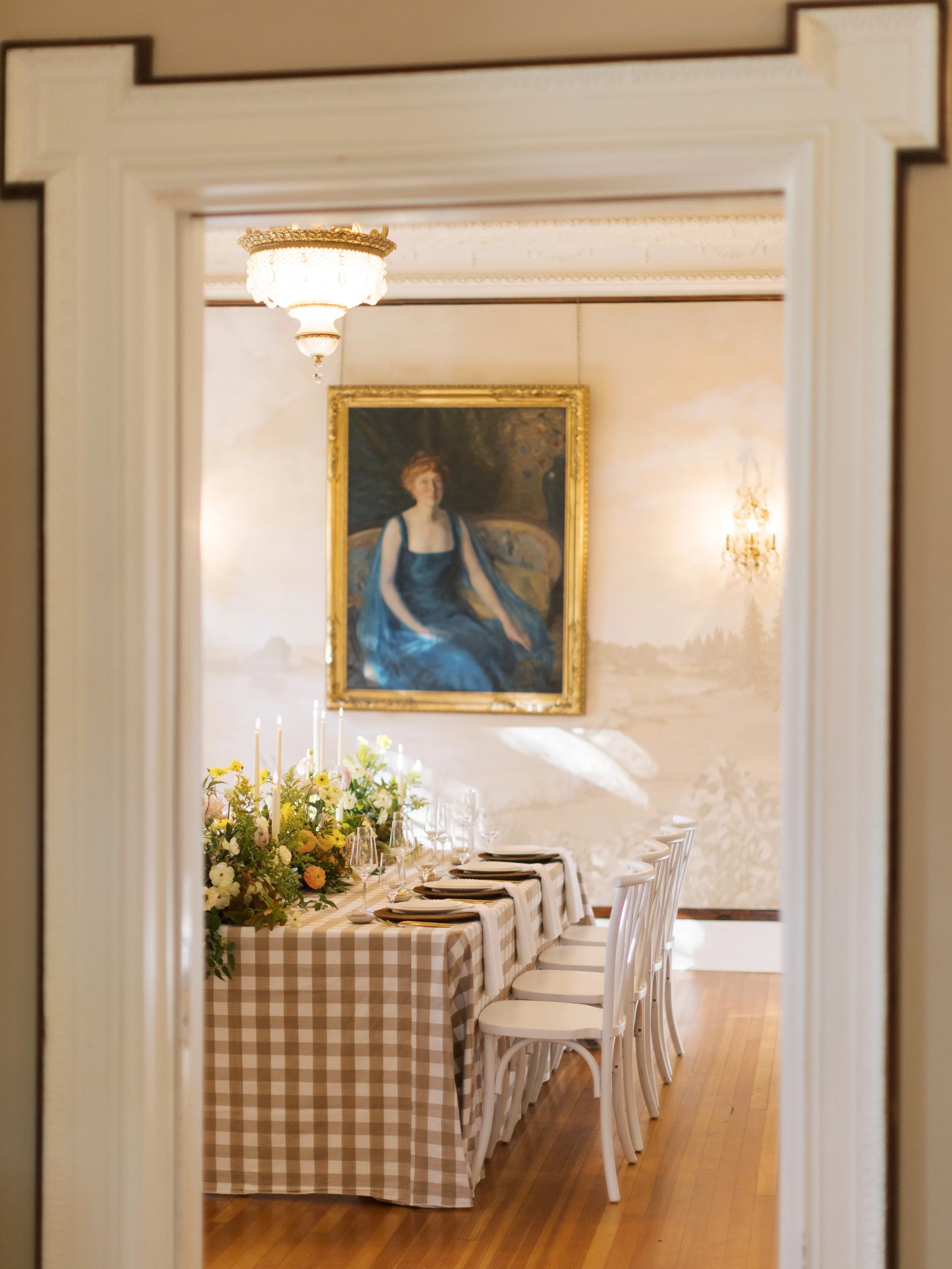 Elegant dining room with a secluded table set with a checkered tablecloth, floral centerpiece, candles, and glassware, viewed through a doorway. A portrait of a woman in a blue dress hangs on the wall, illuminated by chandeliers and wall sconces.
