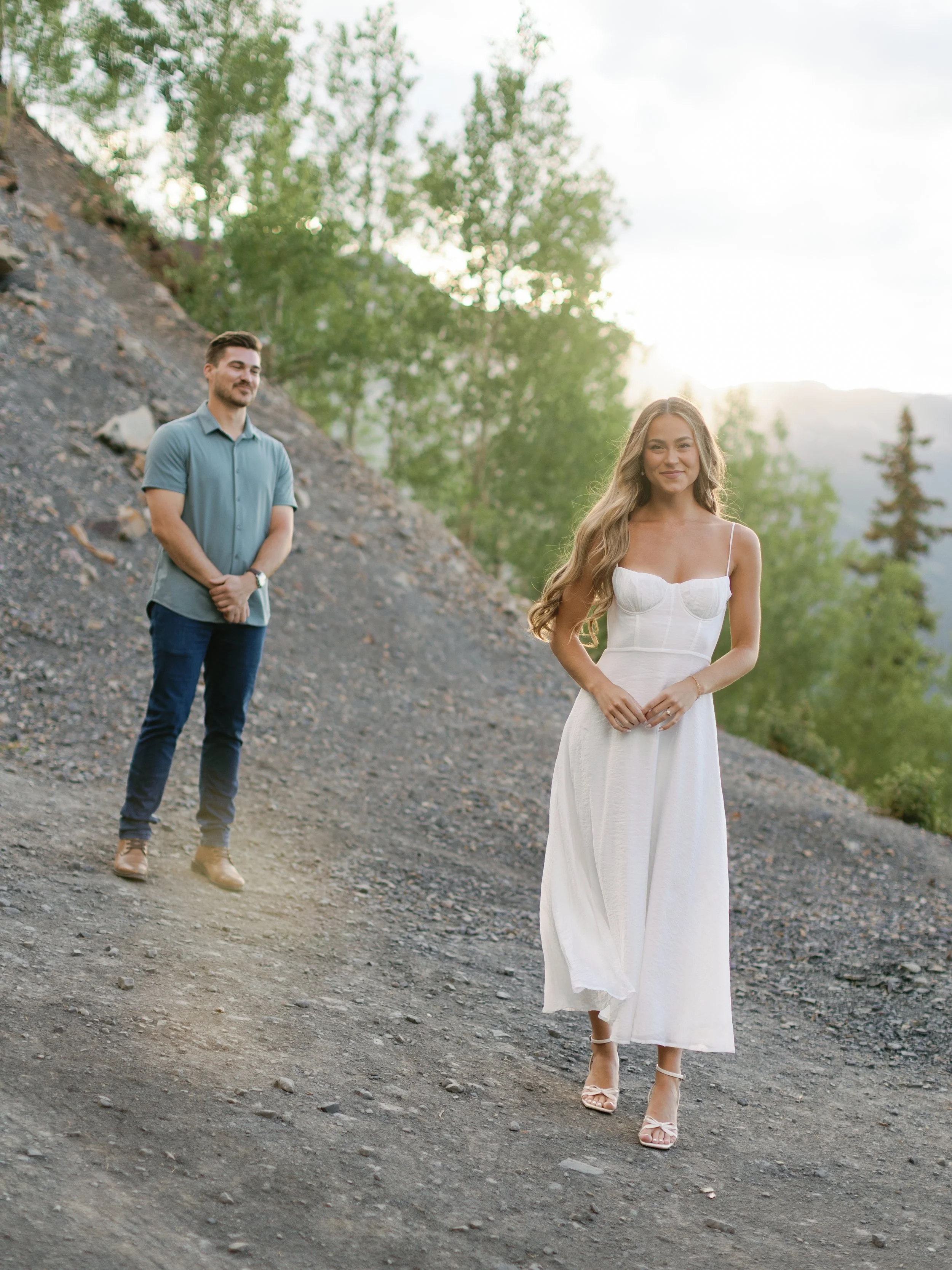 A woman in a white dress walks on rocky terrain with a man standing in the background. The scene is outdoors with trees and a hill, during either sunrise or sunset.
