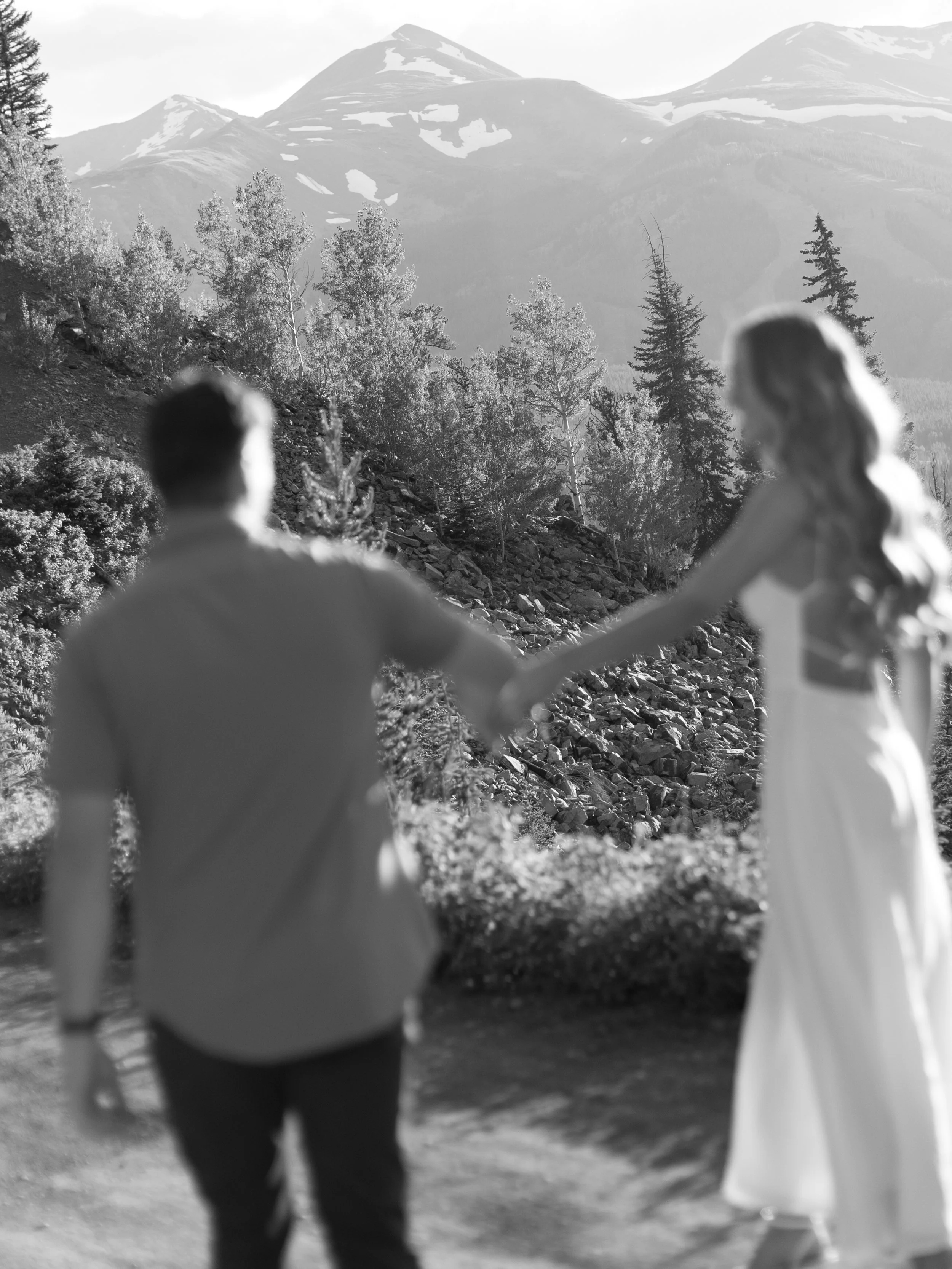 A couple holding hands in a mountainous outdoor setting with trees and snow-capped peaks in the background.