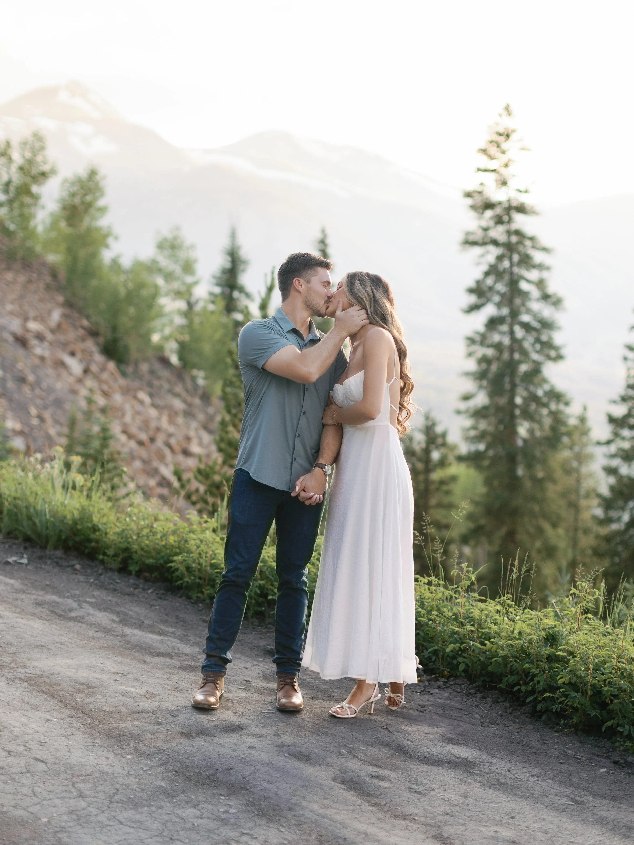A couple kissing outdoors in a scenic mountain setting during daytime, with trees and distant mountains in the background.