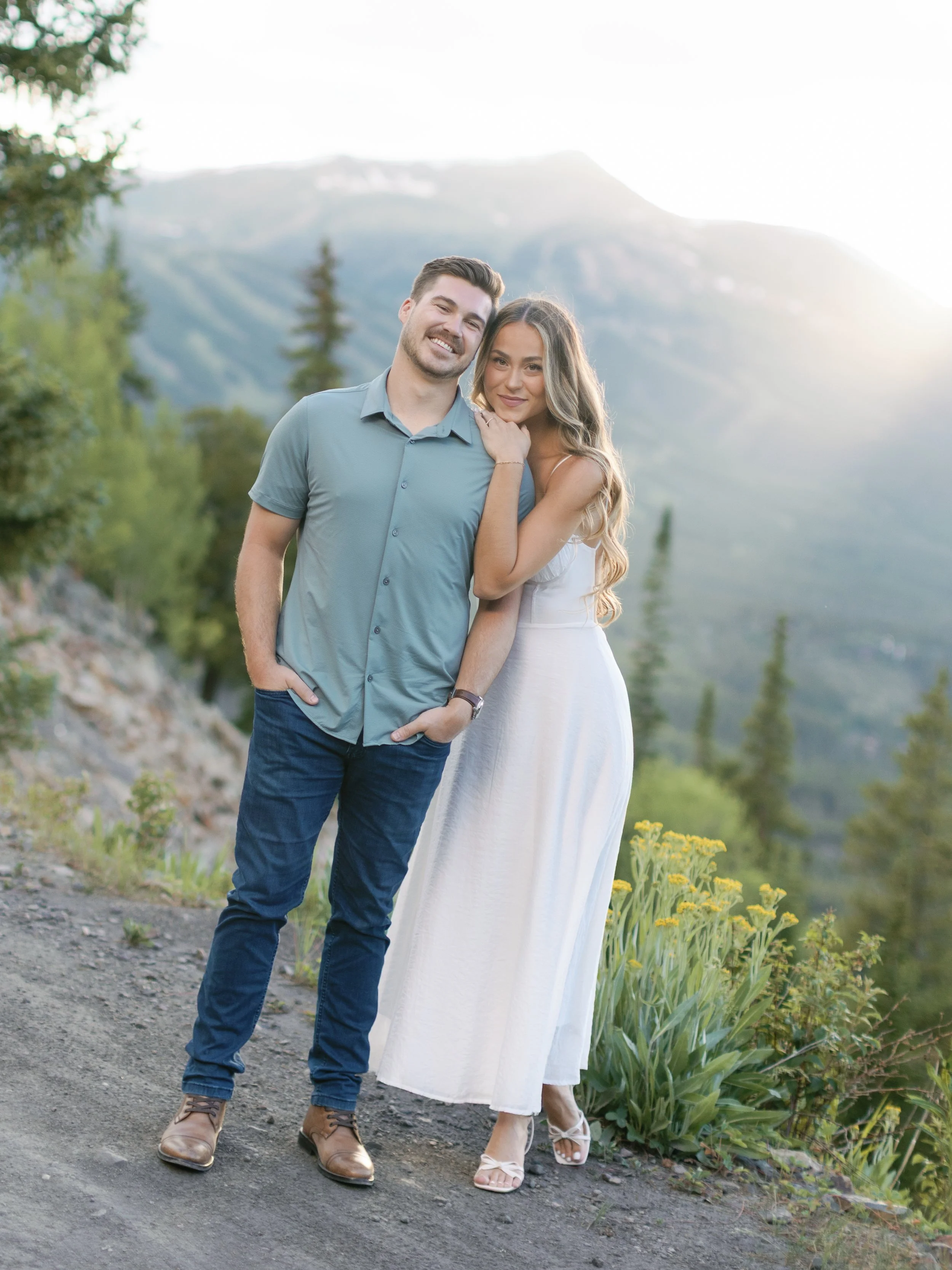 A smiling couple standing outdoors on a dirt path with mountains and trees in the background, during sunset or sunrise.