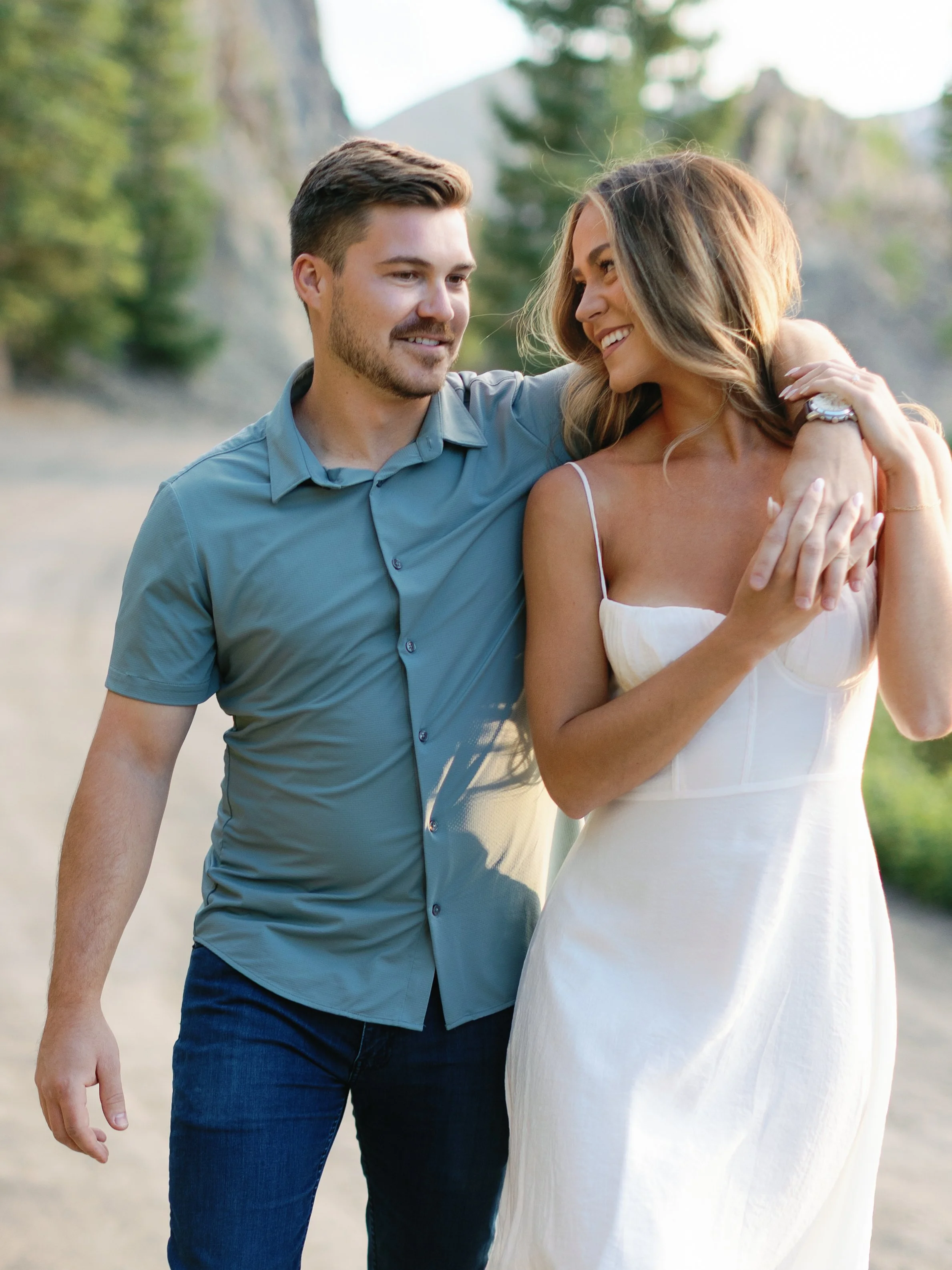A couple walking outdoors, smiling, with the man resting his arm on the woman's shoulder; the woman is wearing a white dress, and the background shows trees and mountains.