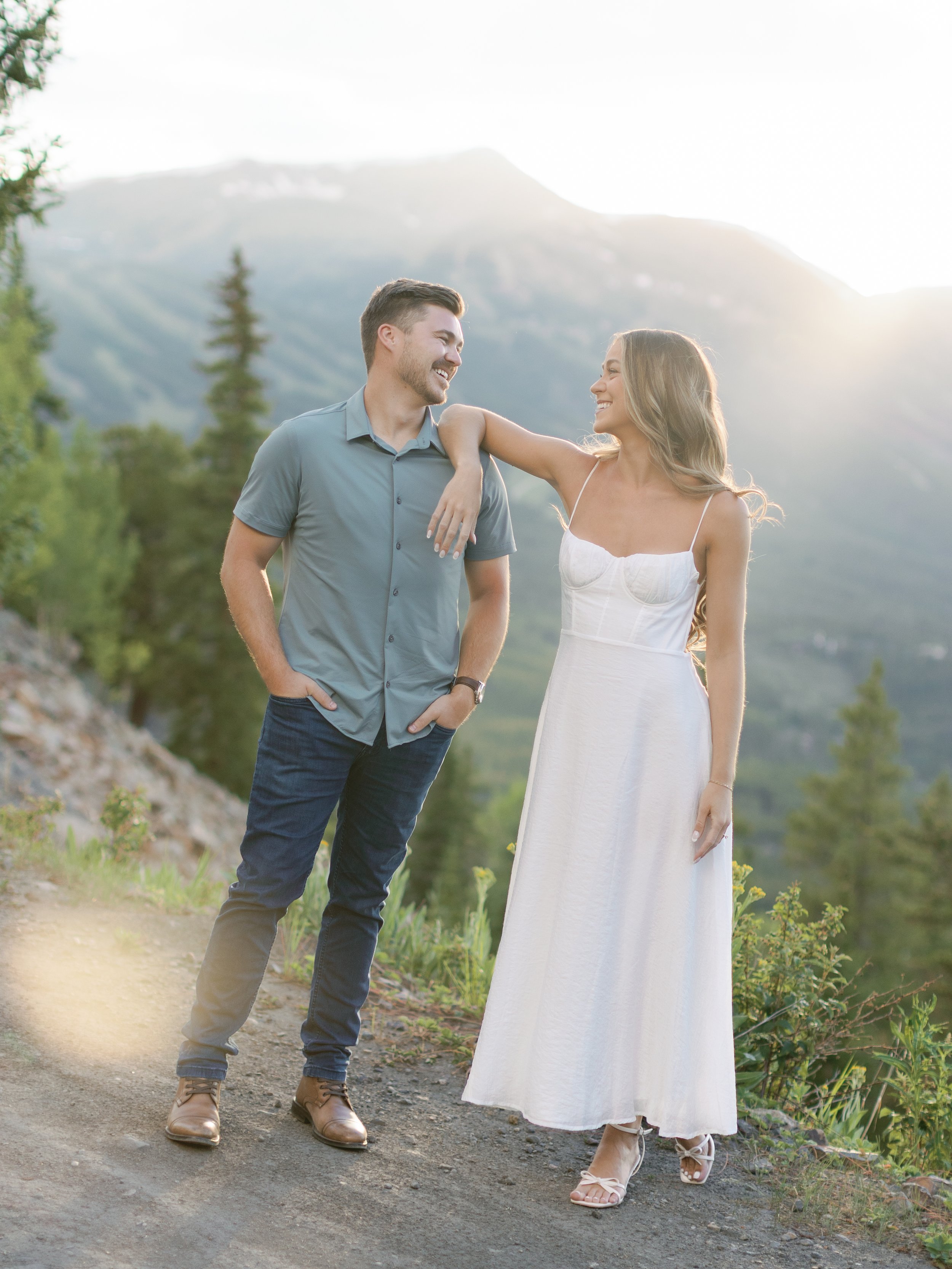 A young couple standing outdoors on a trail with mountains and trees in the background, enjoying each other's company.
