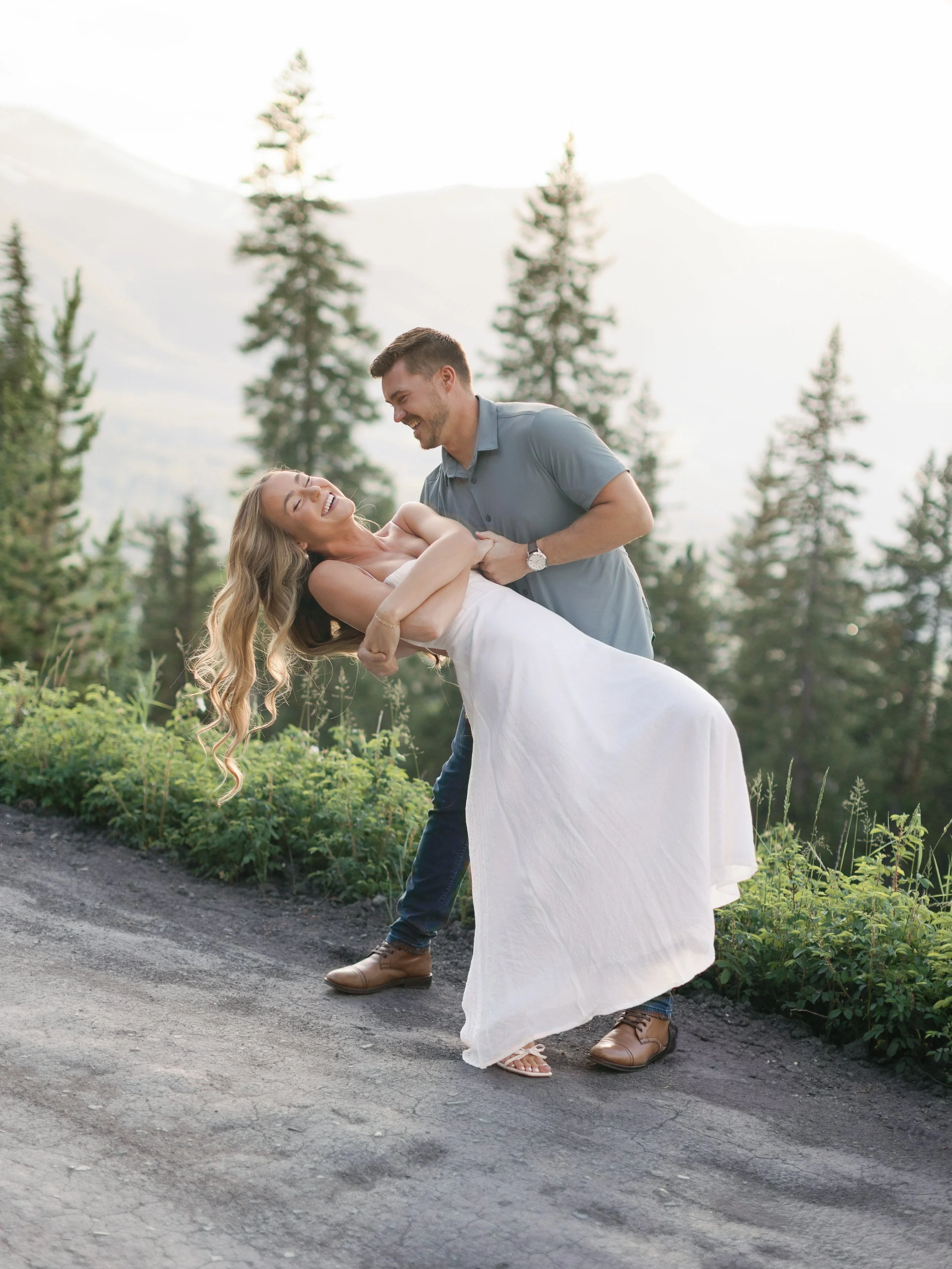 A man and woman are outside on a dirt path surrounded by trees and mountains in the background. The man is holding the woman who is smiling and laughing. The woman wears a white dress, and the man wears a gray shirt and blue jeans.