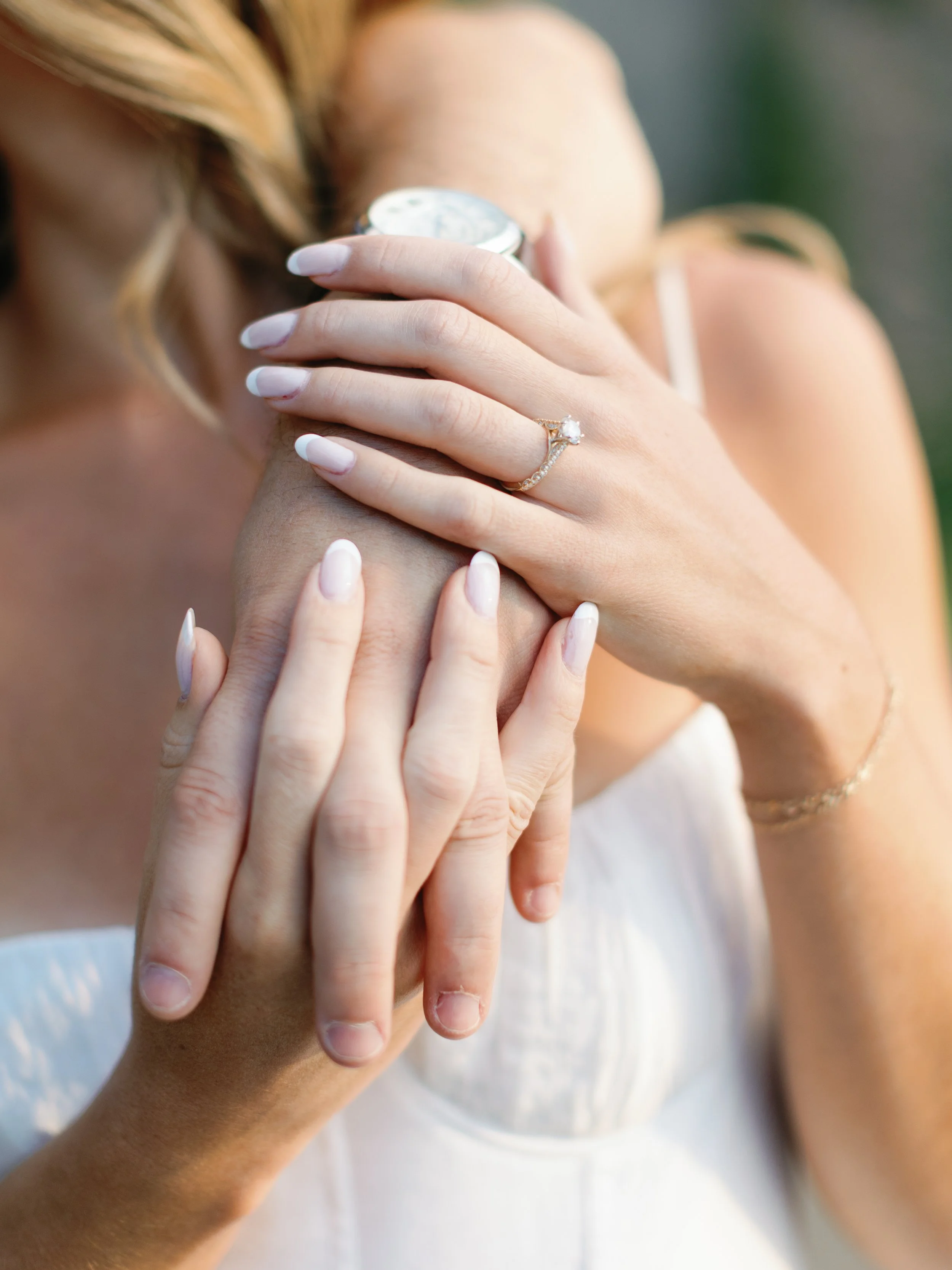 Close-up of a woman showing her wedding ring and engagement ring on her left hand, with her hands resting on her shoulder and forehead, wearing a white dress.