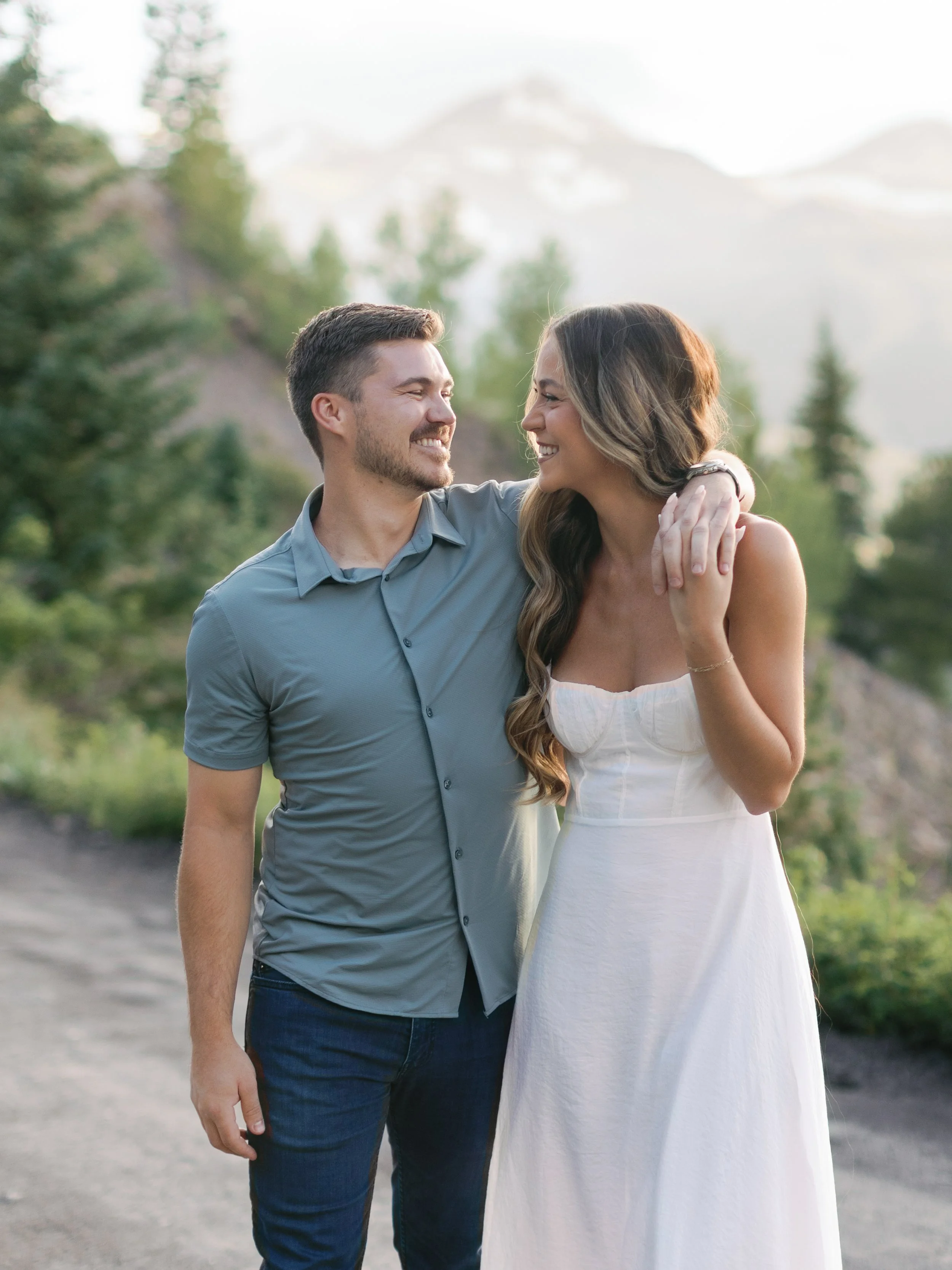 A couple smiling and looking at each other outdoors in a natural setting with mountains and trees in the background.