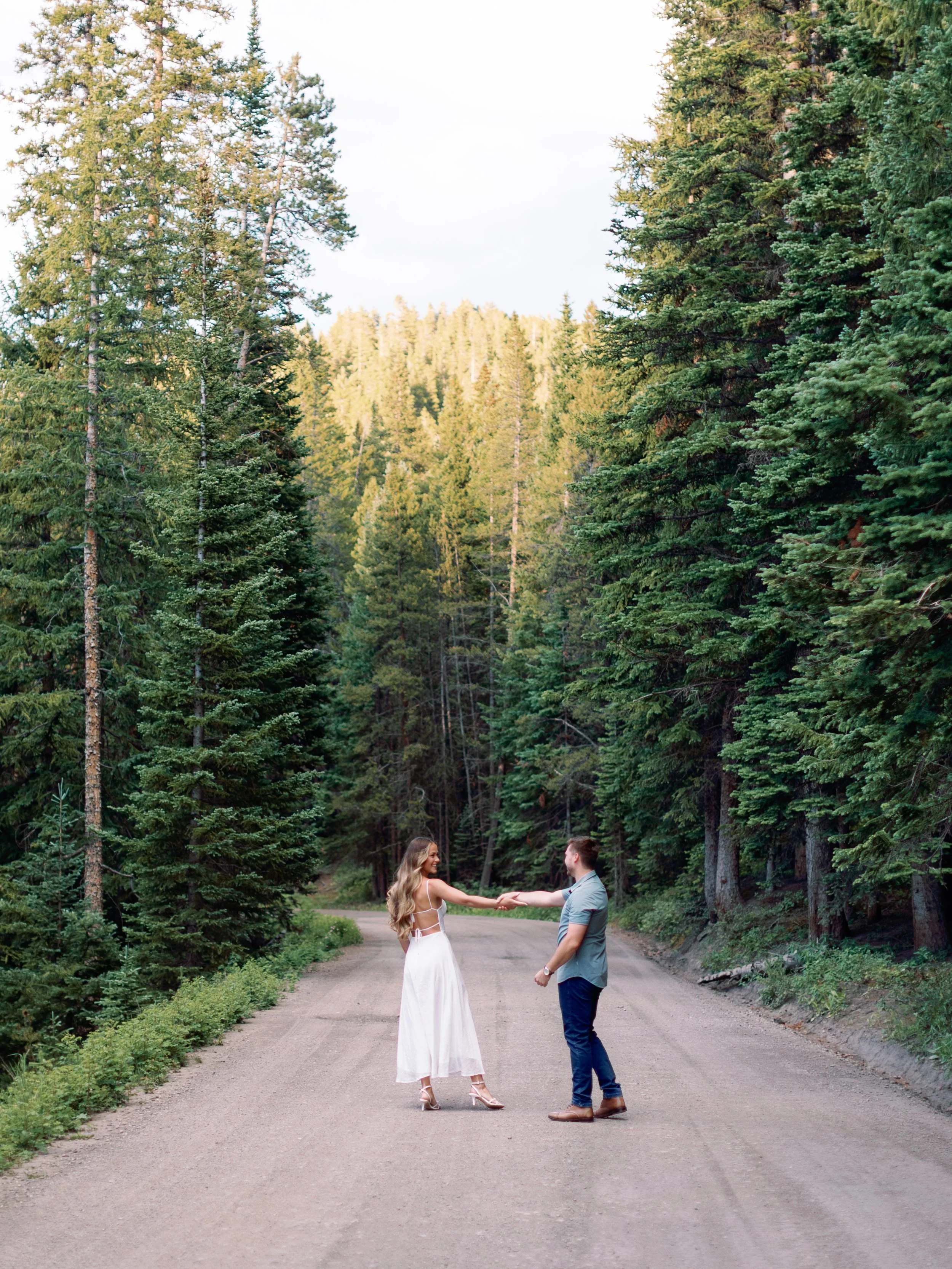 A man and woman holding hands in the middle of a gravel forest road surrounded by tall evergreen trees.