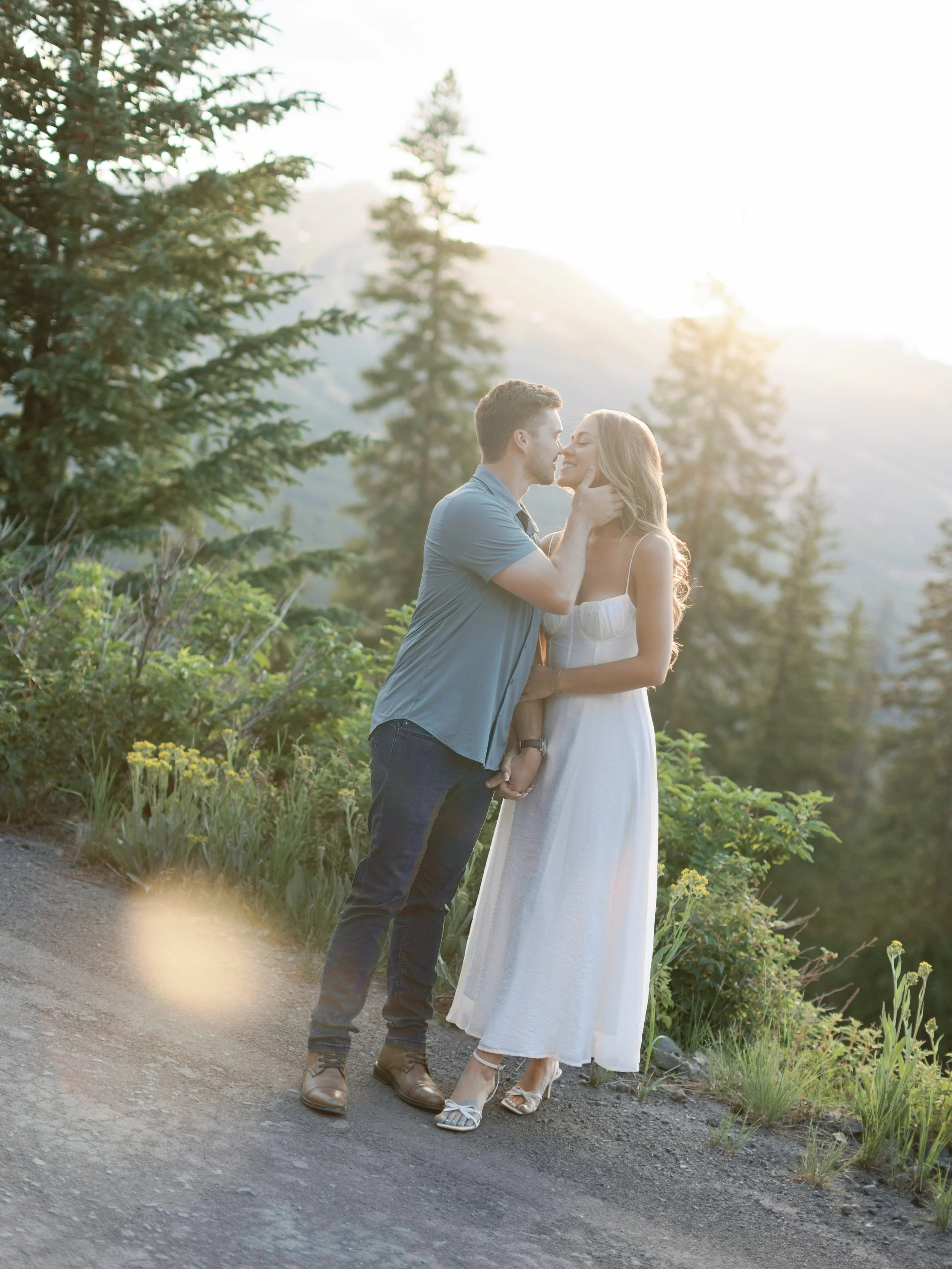 A young couple stands close together outdoors among trees and plants, with the sun setting behind them. The man gently holds the woman's face, and they are about to kiss, sharing a tender moment in a natural, scenic setting.