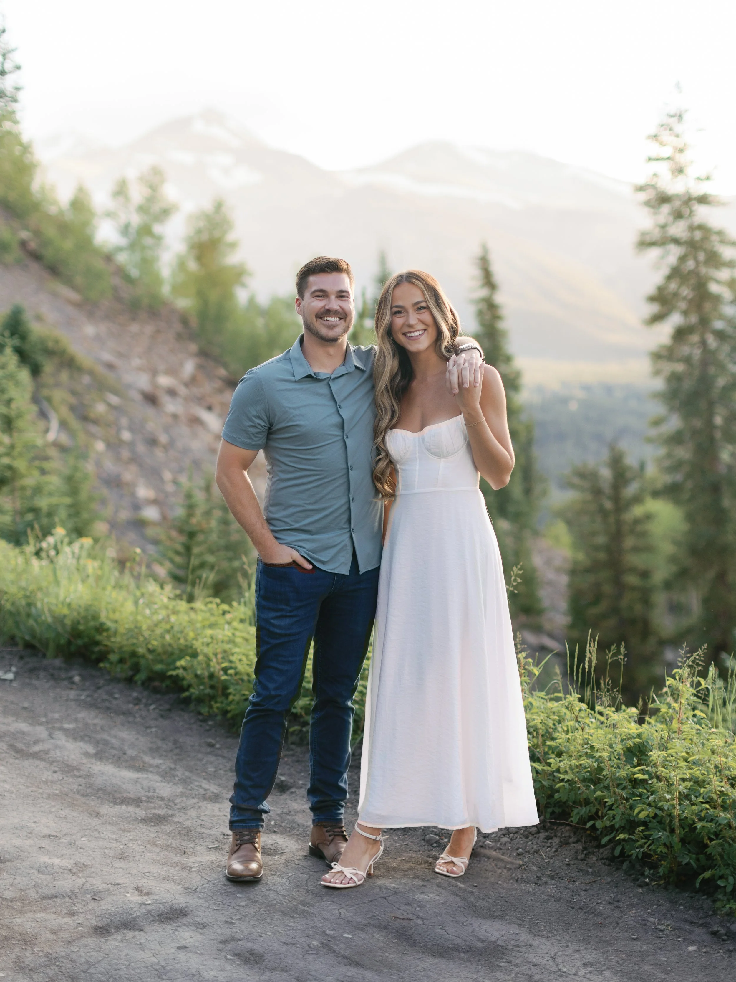 A couple smiling outdoors in a mountain landscape, the woman is wearing a white dress and the man is in a blue shirt and jeans.