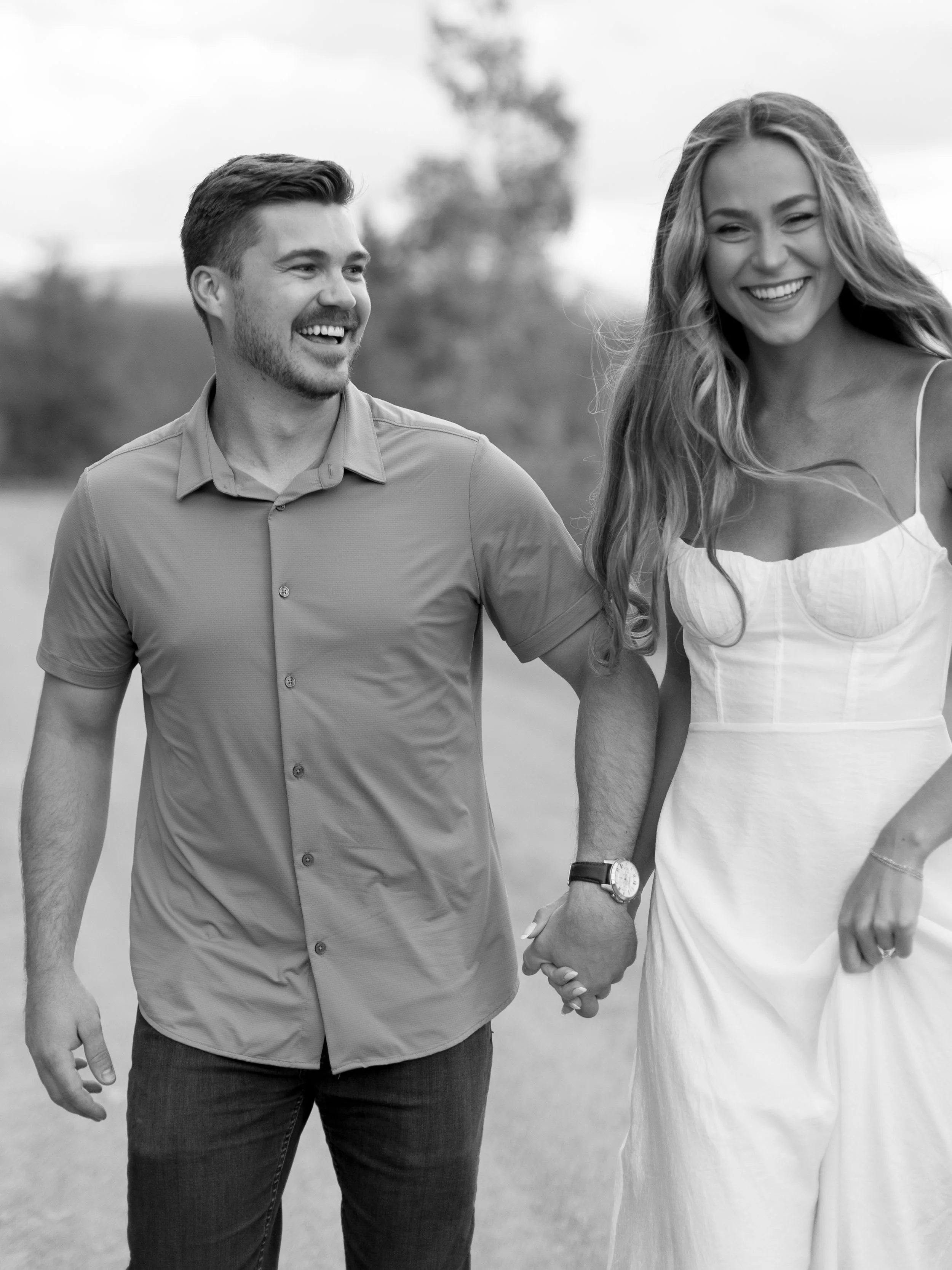 A smiling man and woman holding hands outdoors, both casually dressed, with trees in the background.