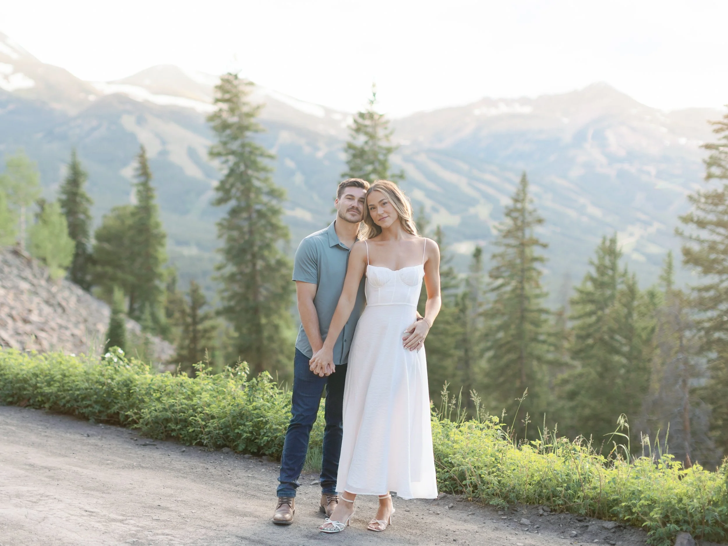 A young couple holding hands, standing outdoors with mountains, trees, and sky in the background.