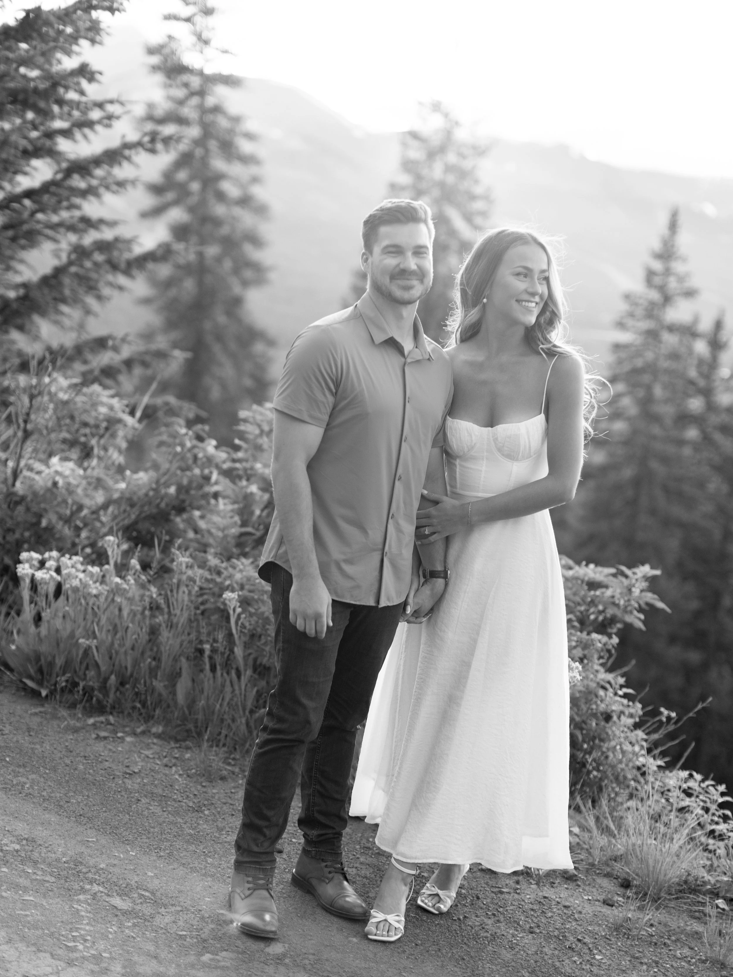 A smiling couple stands outdoors in a scenic mountain and forest setting, holding hands, with the woman wearing a white dress and the man in a button-up shirt and jeans.