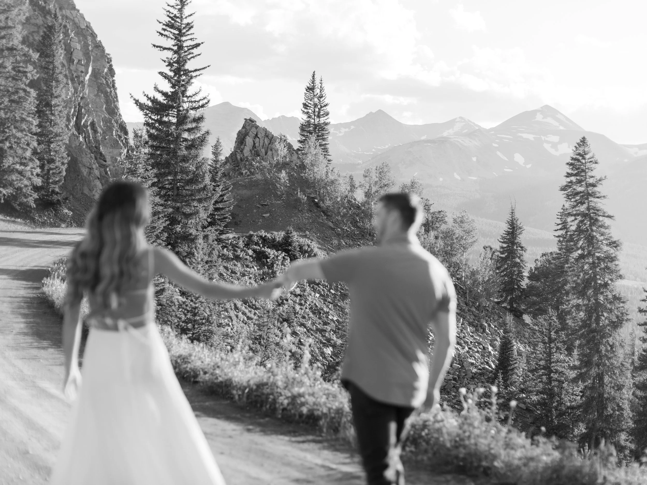 A couple holding hands on a trail in a mountainous forest landscape.