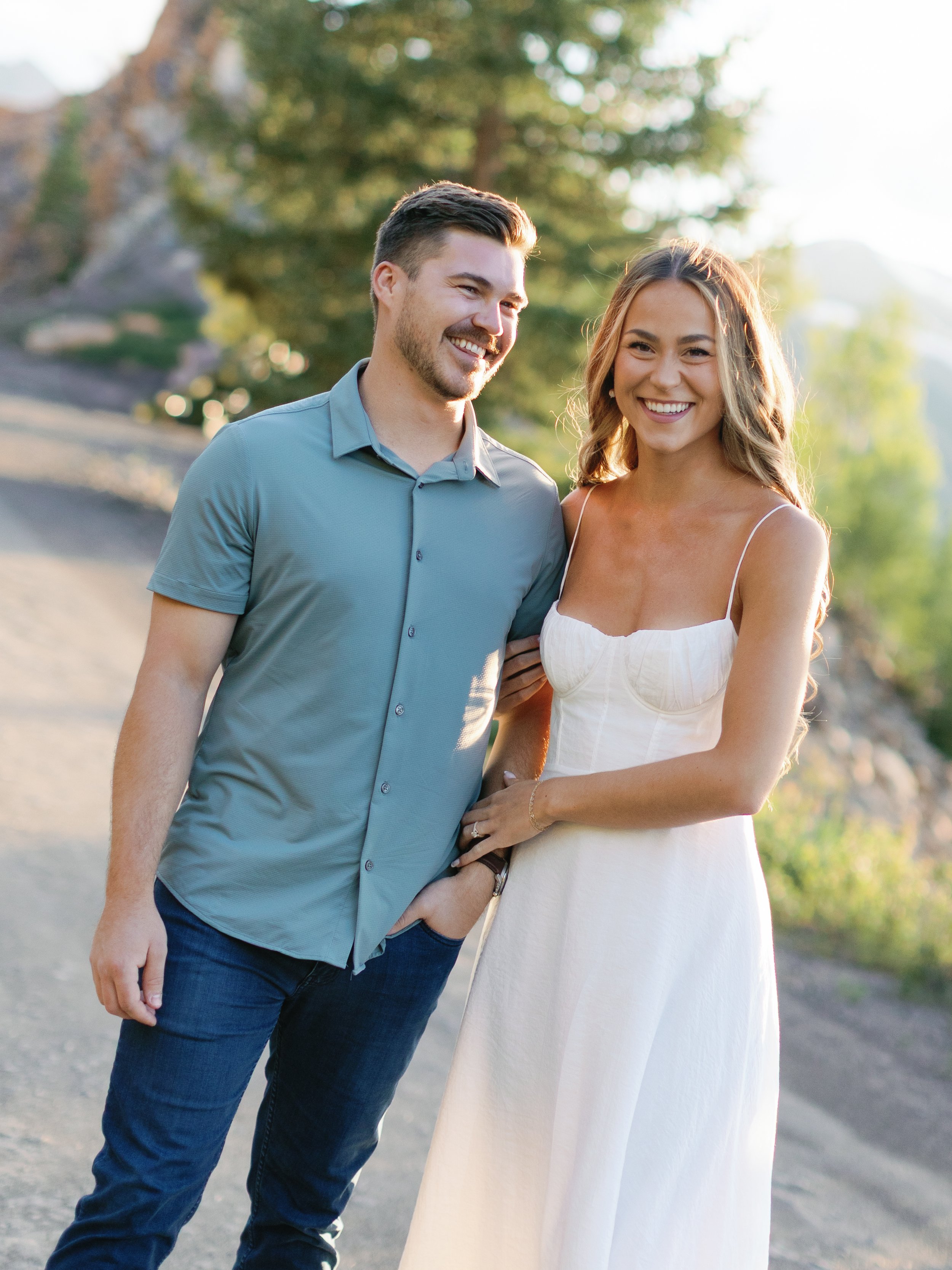 A smiling couple standing outdoors during golden hour, with mountains and trees in the background.