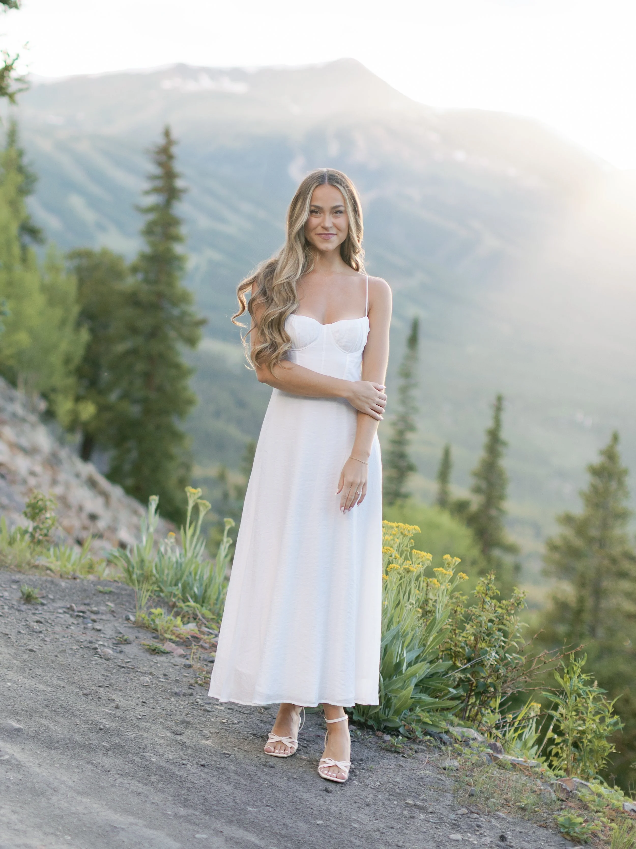 A woman in a white dress standing outdoors on a mountain trail with trees and mountains in the background.