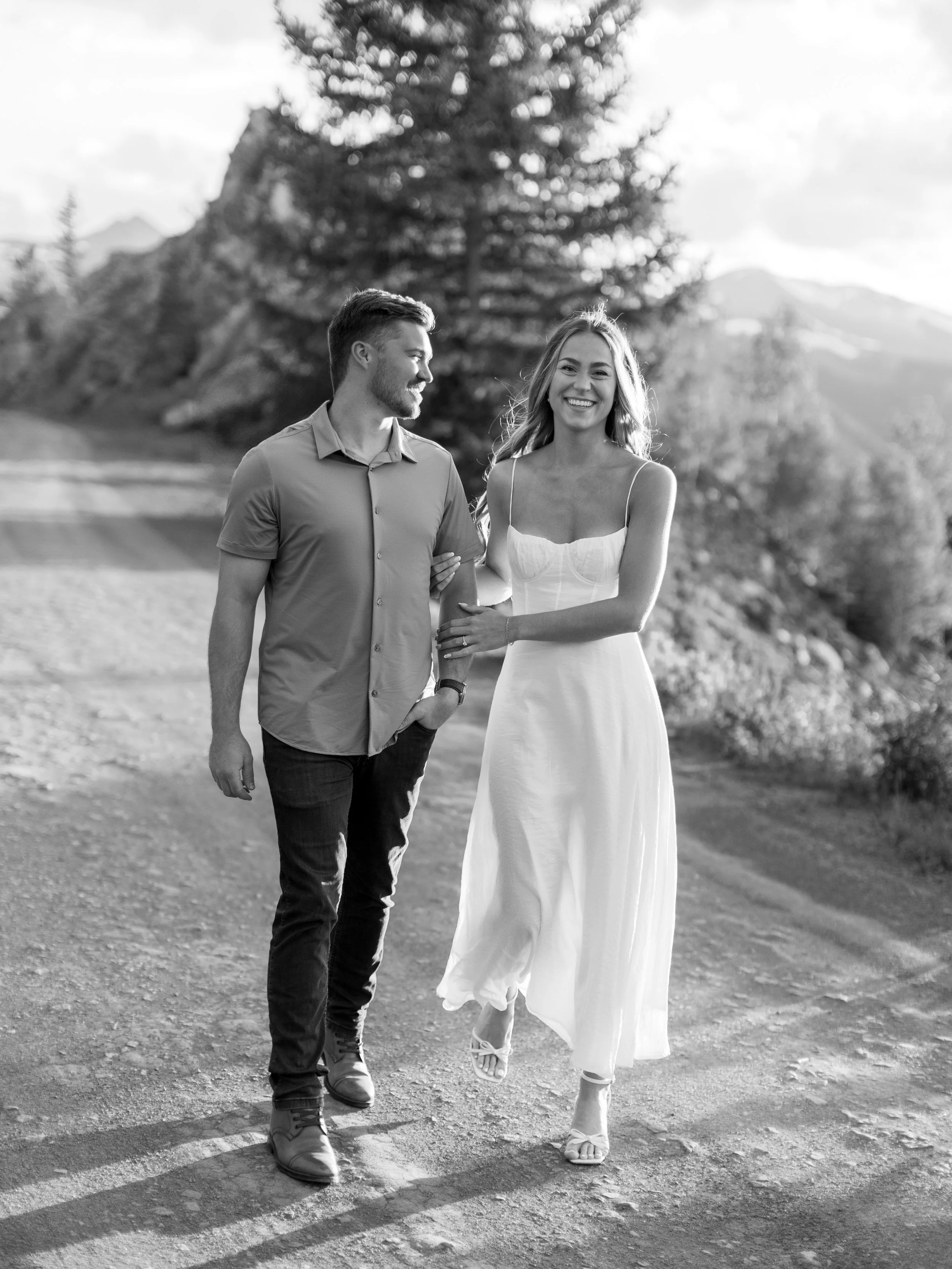 A happy couple walks outdoors on a dirt path, the man in a short-sleeved button-up shirt and dark pants, the woman in a white dress, with mountains and trees in the background, captured in black and white.